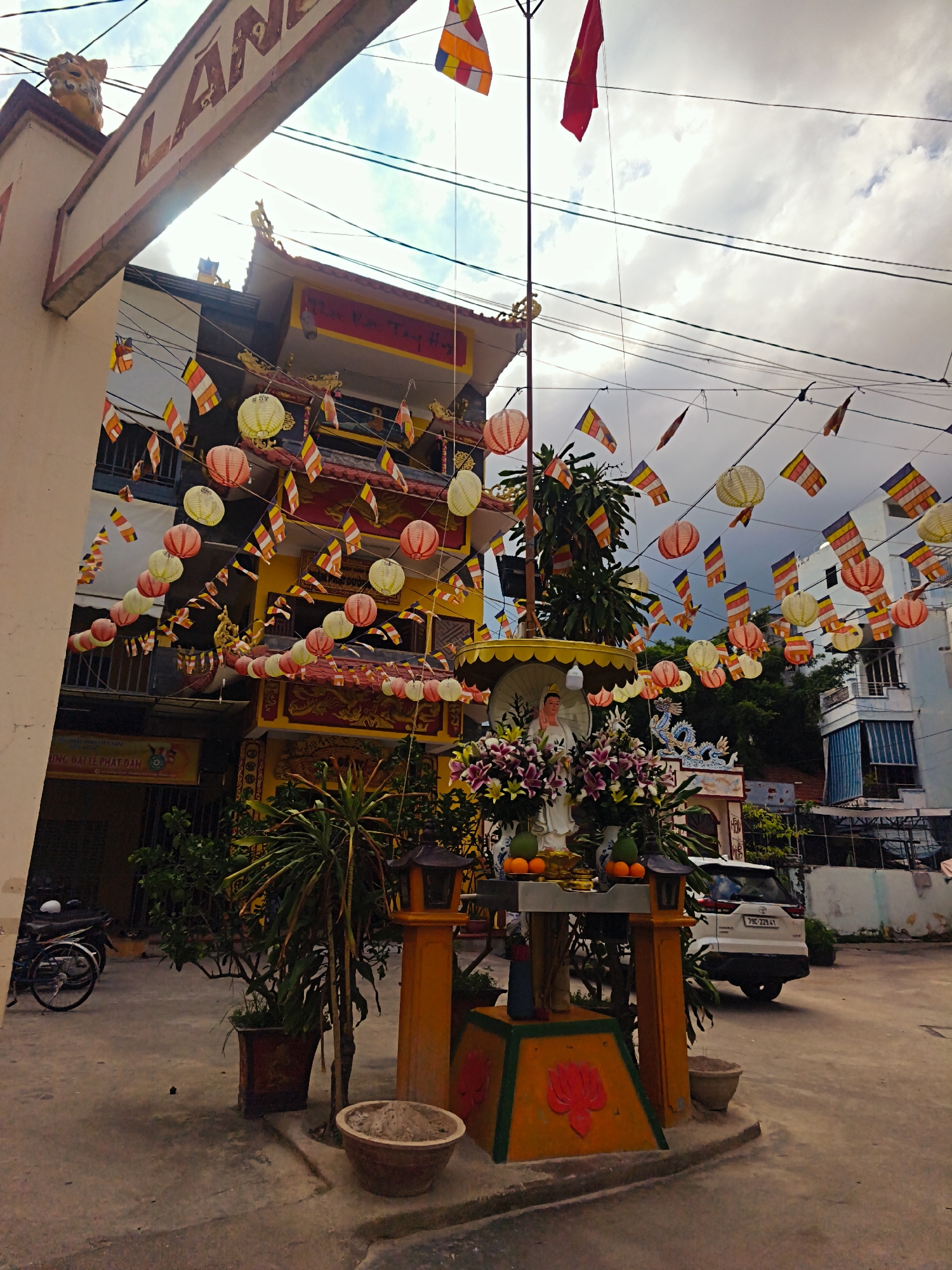 This image captures a street view in Nha Trang, Vietnam, likely near a temple or cultural site. The foreground features a paved courtyard with several potted plants and a decorative altar displaying a statue of a deity surrounded by flowers and fruit. To the right, a white SUV is parked. The background is dominated by a multi-story building with ornate architectural details, adorned with numerous colorful flags and lanterns strung across wires. Several motorcycles are visible parked to the left of the building. The sky is overcast, with visible power lines crisscrossing the scene. The overall impression is one of a vibrant, culturally rich urban setting.