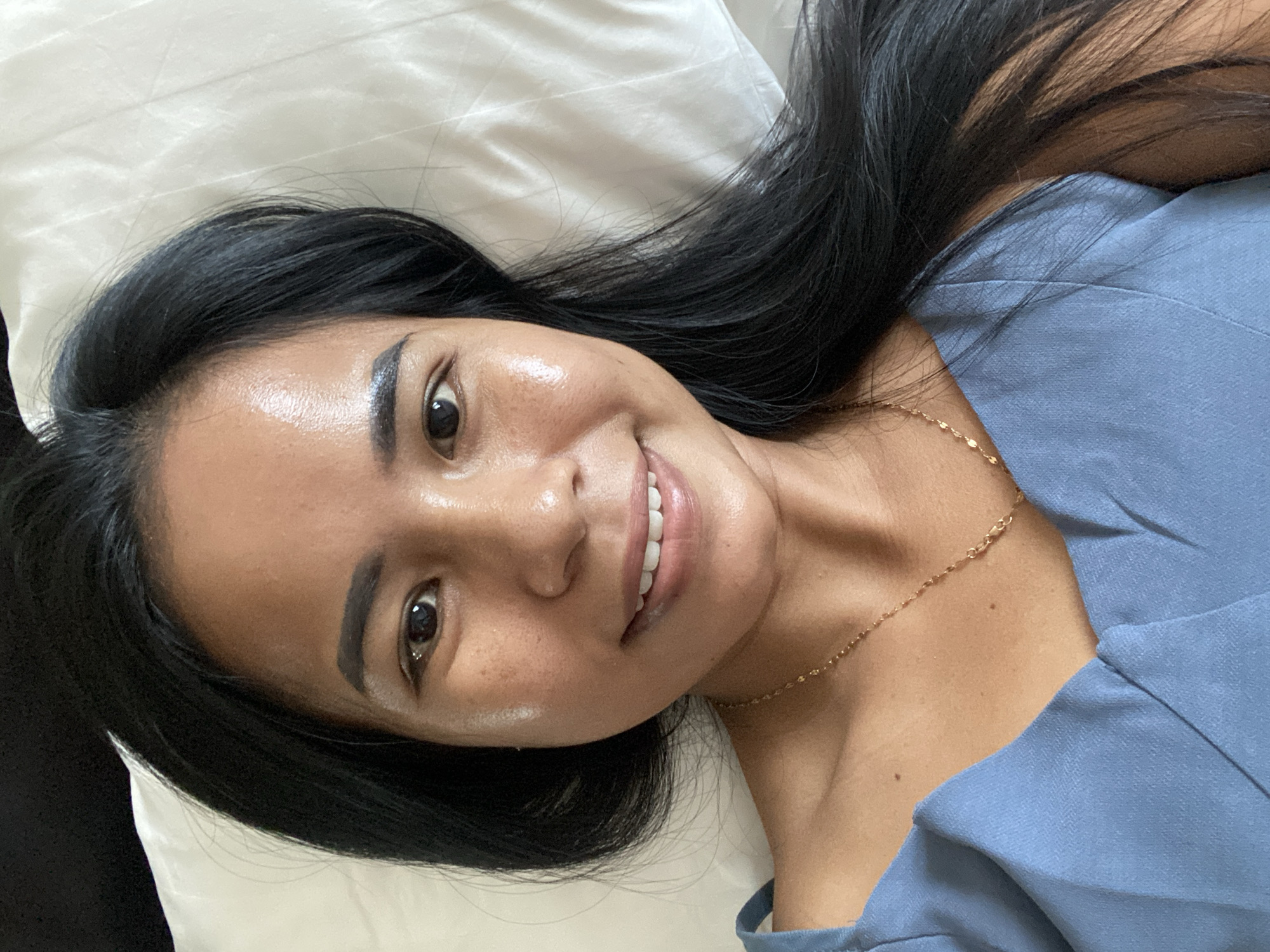 The image shows a smiling Southeast Asian woman lying on a white pillow. She has long black hair, brown eyes, and is wearing a light blue top and a gold chain necklace. Her location cannot be determined solely from this image.