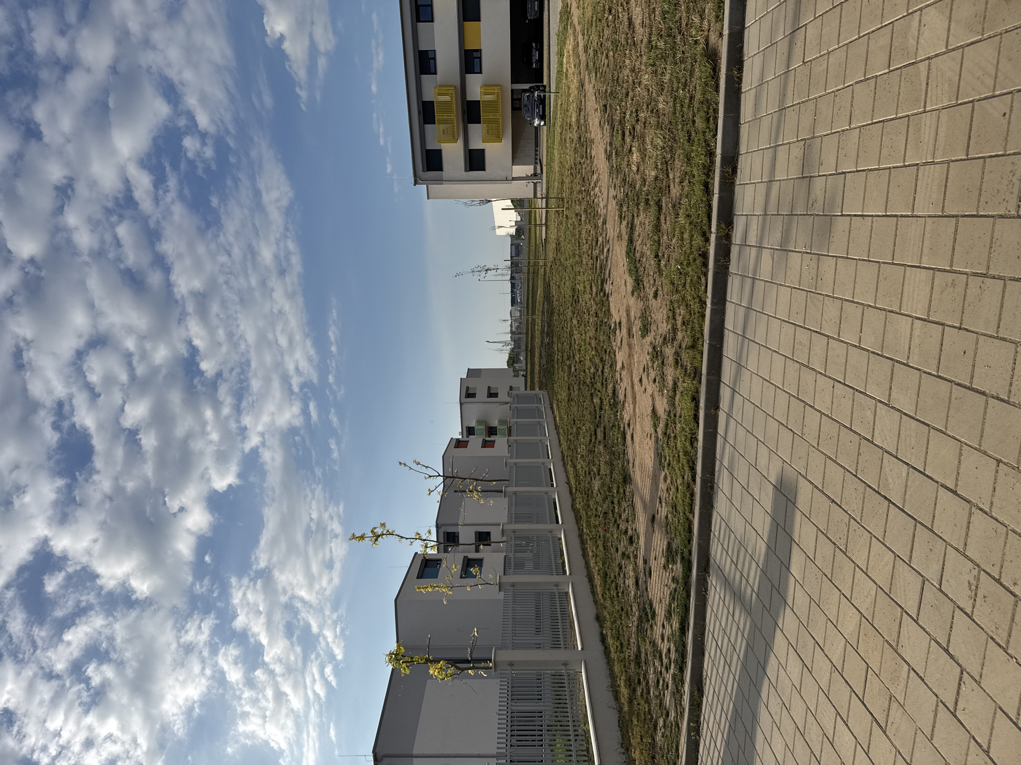 The image depicts a modern residential area under a partly cloudy sky. In the foreground, a paved walkway made of light-colored rectangular bricks runs diagonally from the bottom left to the bottom right. Adjacent to the walkway is a strip of dry, sparse grass bordered by a concrete curb. Behind the grass is a metal fence with vertical slats supported by concrete posts, separating the walkway area from the buildings.
Beyond the fence, a row of contemporary, multi-story apartment buildings with white and gray facades is visible. These buildings have dark-framed windows and some feature bright yellow balconies or accents. Young trees with sparse foliage are planted along the front of the buildings, between the fence and the structures.
Further in the background, more buildings and a glimpse of distant industrial structures can be seen. Several cars are parked in front of one of the apartment buildings, including a dark sedan and a black SUV. The overall scene suggests a newly developed or developing urban residential zone with a focus on modern architecture and landscaping.