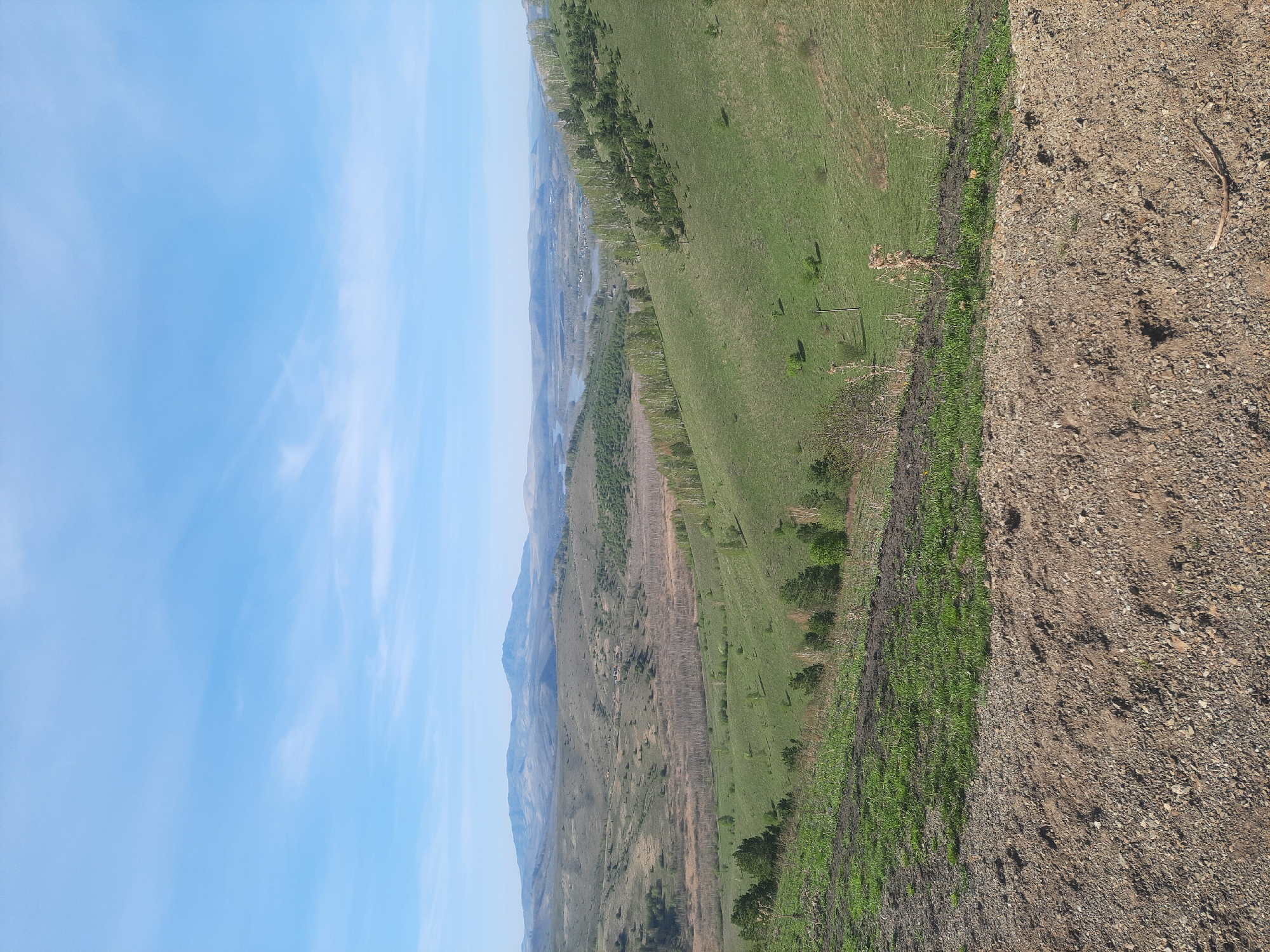 This media file captures a wide, expansive landscape view, likely from an elevated position overlooking a valley. The scene is dominated by natural elements, suggesting a rural or mountainous region in Aya, Russia.
In the foreground, to the right, there is a rough, unpaved area composed of dirt and gravel, possibly a track or a roadside embankment. This transitions into a lush green hillside, covered in grass and dotted with scattered trees and shrubs.
The midground reveals a vast valley. A prominent feature is a winding body of water, likely a river or lake, that stretches into the distance. Along the banks of this water body, and across the valley floor, are more green fields and numerous trees, some appearing as dense rows or groves. In the far midground, near the water, faint structures suggest a small settlement or village nestled within the landscape. Beyond these, rolling hills and mountains rise, their outlines softened by distance and atmospheric haze.
The background consists of a bright, clear blue sky with delicate, wispy white clouds trailing across it, indicating fine weather. The time of day appears to be bright daylight, possibly morning or early afternoon, with even illumination across the landscape.
There are no people or discernible activities taking place, and no visible text in the image. The overall impression is one of natural serenity and vastness.