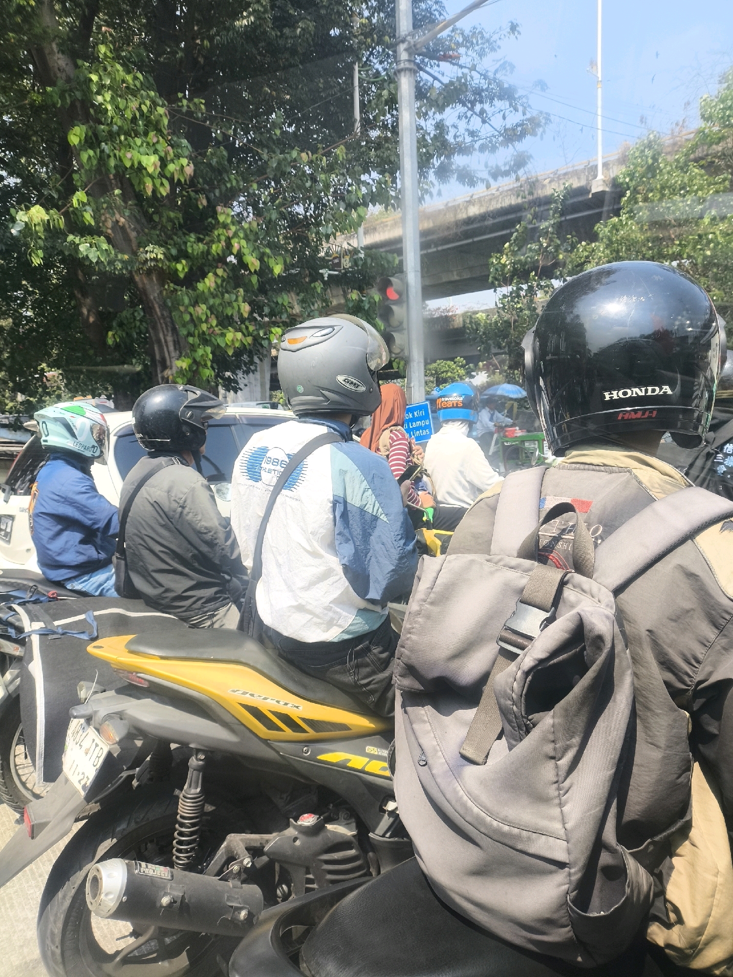 The image depicts a traffic scene on a sunny day in West Jakarta, Indonesia. Several motorcycles are visible, with riders wearing helmets. In the foreground, a person wearing a black helmet and a grey backpack is seated on a yellow and black motorcycle. Another person in a grey helmet and a white and blue jacket is in front of them. To the left, a person in a blue jacket and a light blue helmet is also on a motorcycle. In the background, there are cars, trees, and an elevated road structure. A traffic light is visible with a red signal illuminated. The overall impression is a typical urban traffic jam with various vehicles and pedestrians.