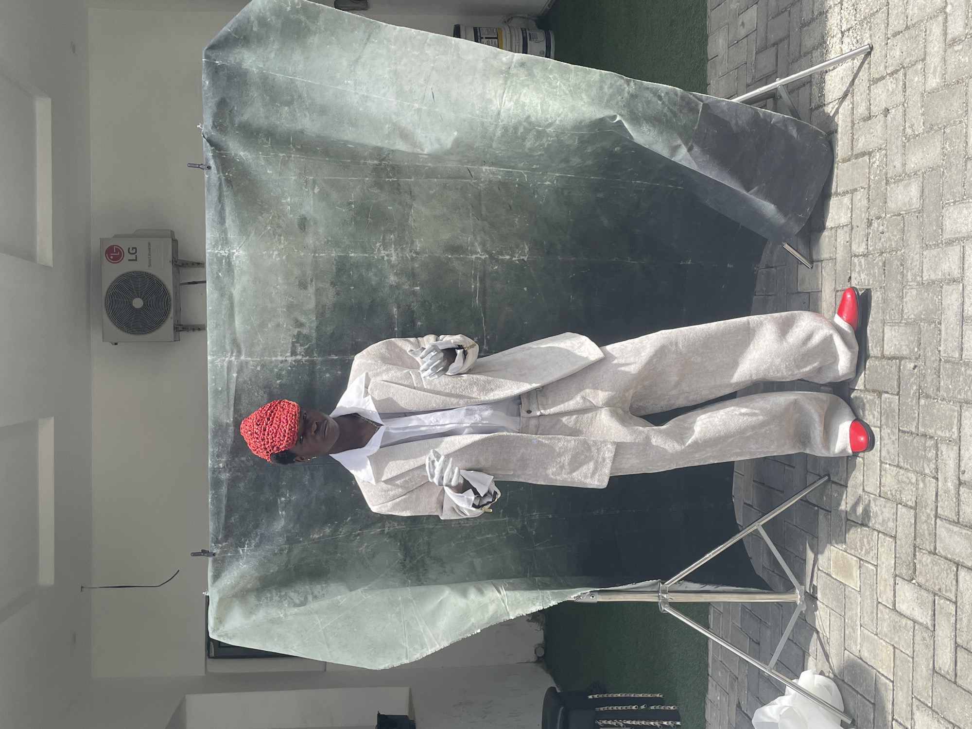 A full-body shot shows a person in a light-colored suit standing in front of a backdrop held up by a support. The person is wearing a red knit cap and red shoes, a white shirt under a light-colored jacket, and matching pants. The person is holding white gloves in both hands. The backdrop is a faded green color. The background shows a building with an LG air conditioning unit mounted on the wall. The scene is on a paved surface that appears to be a patio, with a patch of green artificial turf visible. Given the architectural style and the LG air conditioning unit visible on the building, this scene is plausibly located in Lekki, Nigeria, although this cannot be confirmed with absolute certainty from the visual data alone.