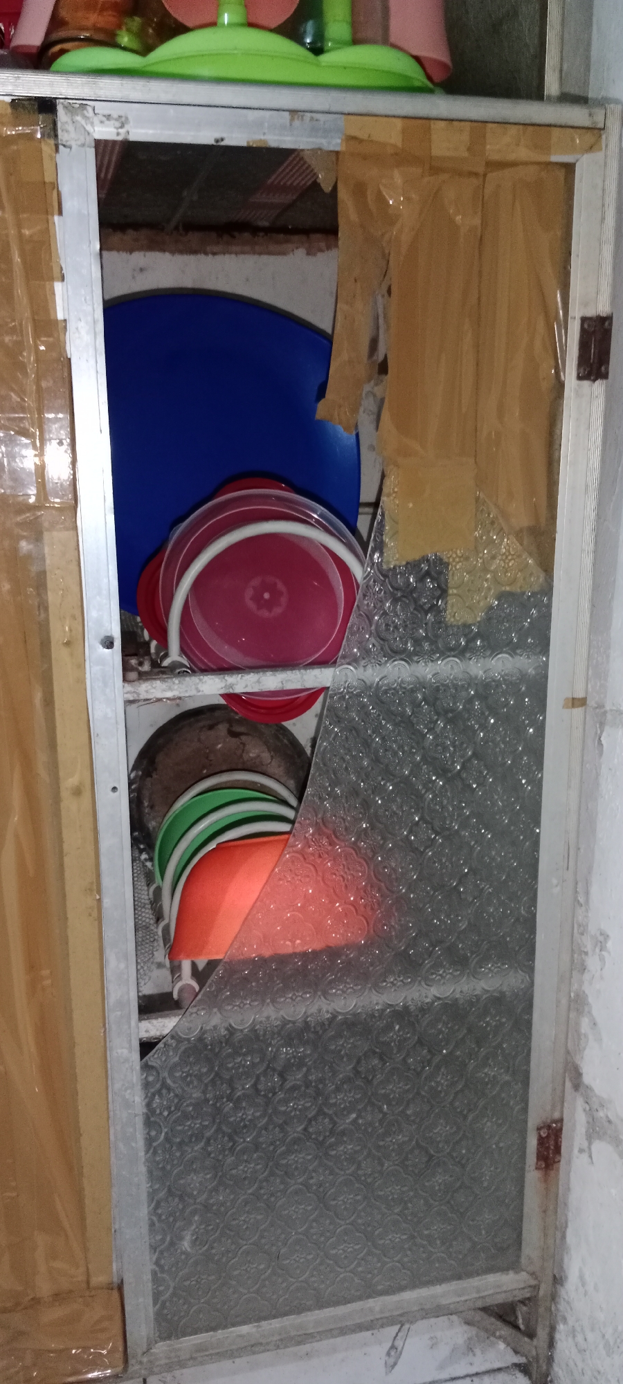 The image shows an open cabinet with several stacked plastic buckets. A large blue bucket is at the top, followed by red buckets, and then green and orange buckets at the bottom. The cabinet door is broken and partially covered with a textured glass panel and taped brown paper. The scene appears to be in a poorly maintained interior space. The location is identified as Subang, Indonesia.