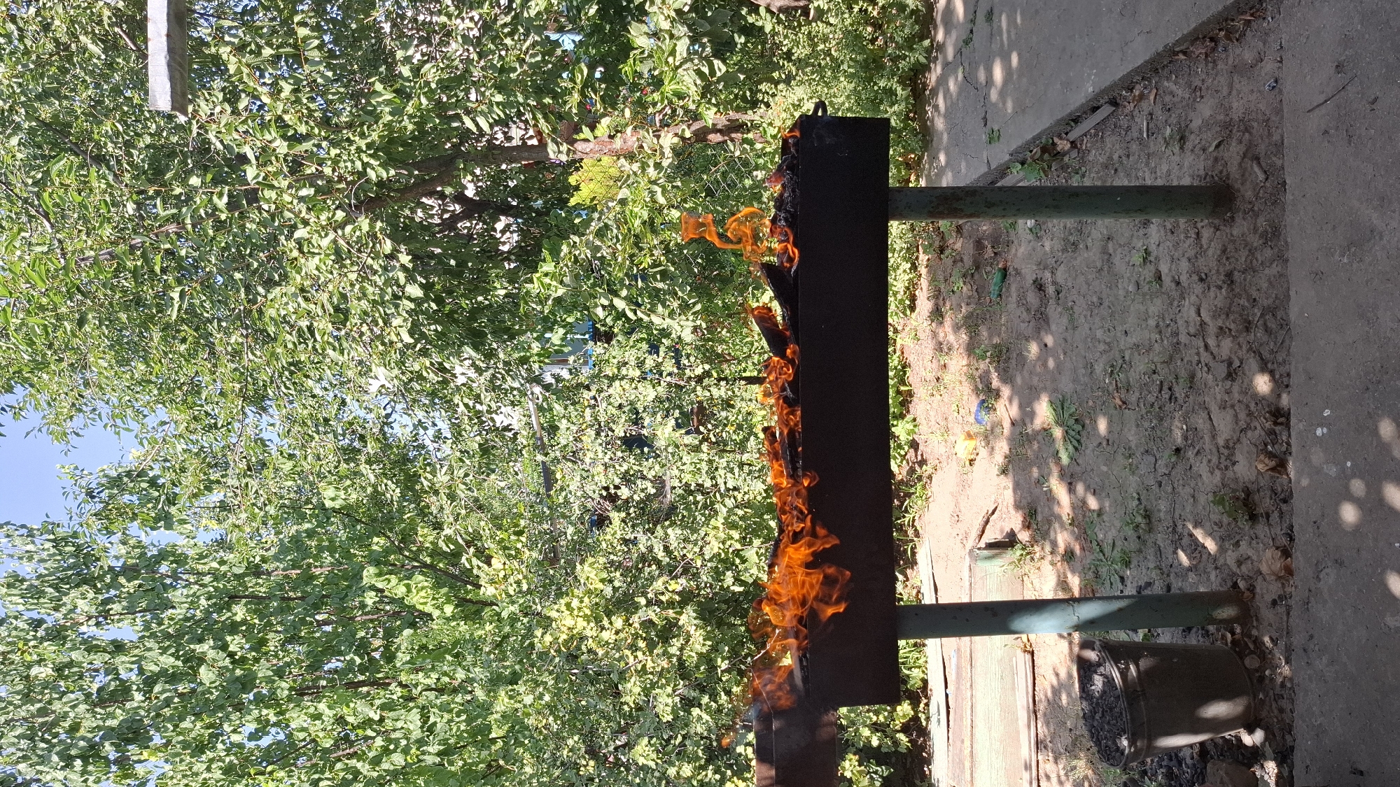 A black metal barbecue grill, filled with burning wood, sits on a green metal stand in an outdoor setting. The grill is positioned on a dirt ground area, with a concrete path to the right and a metal bucket filled with ash to the left. Lush green trees and foliage surround the scene, with a clear blue sky visible above. The overall impression is that of a rustic, possibly residential, backyard. The location is specified as Shubaršu, Kazakhstan. There are no people visible in the image. The scene is calm and still, with the focus being on the barbecue and its surrounding environment.