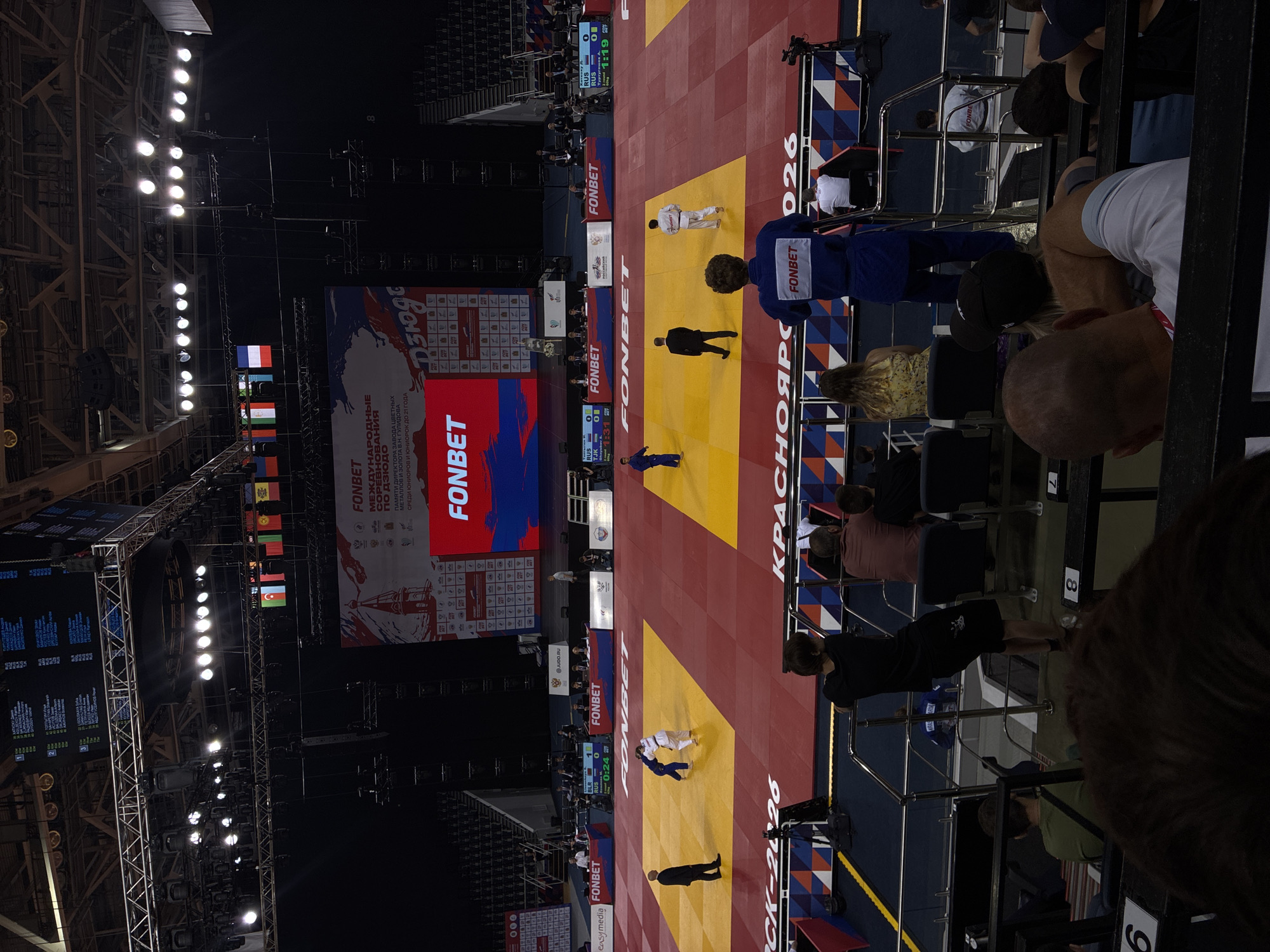 An indoor judo competition is underway in an arena. Several judokas in white and blue gis are on the mat, with two actively engaged in a match while others stand by. A referee in a suit observes the action. Spectators are seated in tiered seating in the foreground, some looking towards the mat, others facing away. A large screen at the back of the arena displays event information and logos, including "FONBET". Flags of various countries are hung across the upper section of the arena. The lighting is bright, illuminating the competition area. The overall scene depicts a professional sporting event in progress.