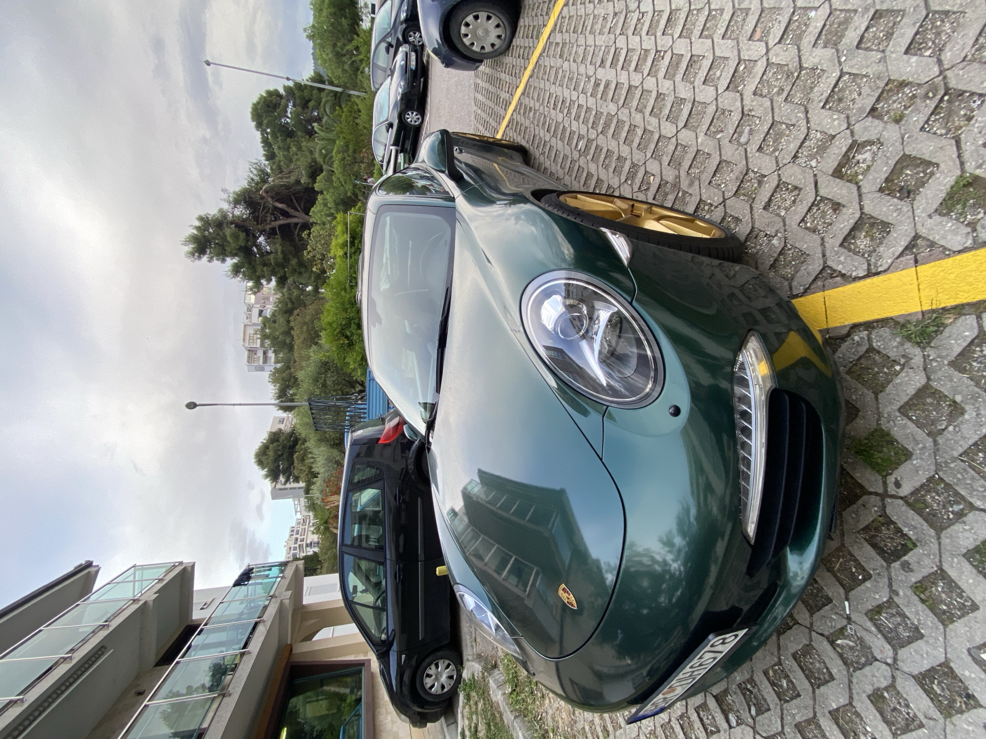 A green Porsche is parked on a paved parking lot in Bar, Montenegro. The front of the car is prominently visible, with the license plate partially obscured. The car is positioned next to a yellow parking line. A black minivan and other vehicles are parked behind the Porsche. In the background, there are trees, buildings, and a cloudy sky. A street lamp is also visible. The parking lot pavement is composed of interlocking concrete blocks with some grass growing in the seams.