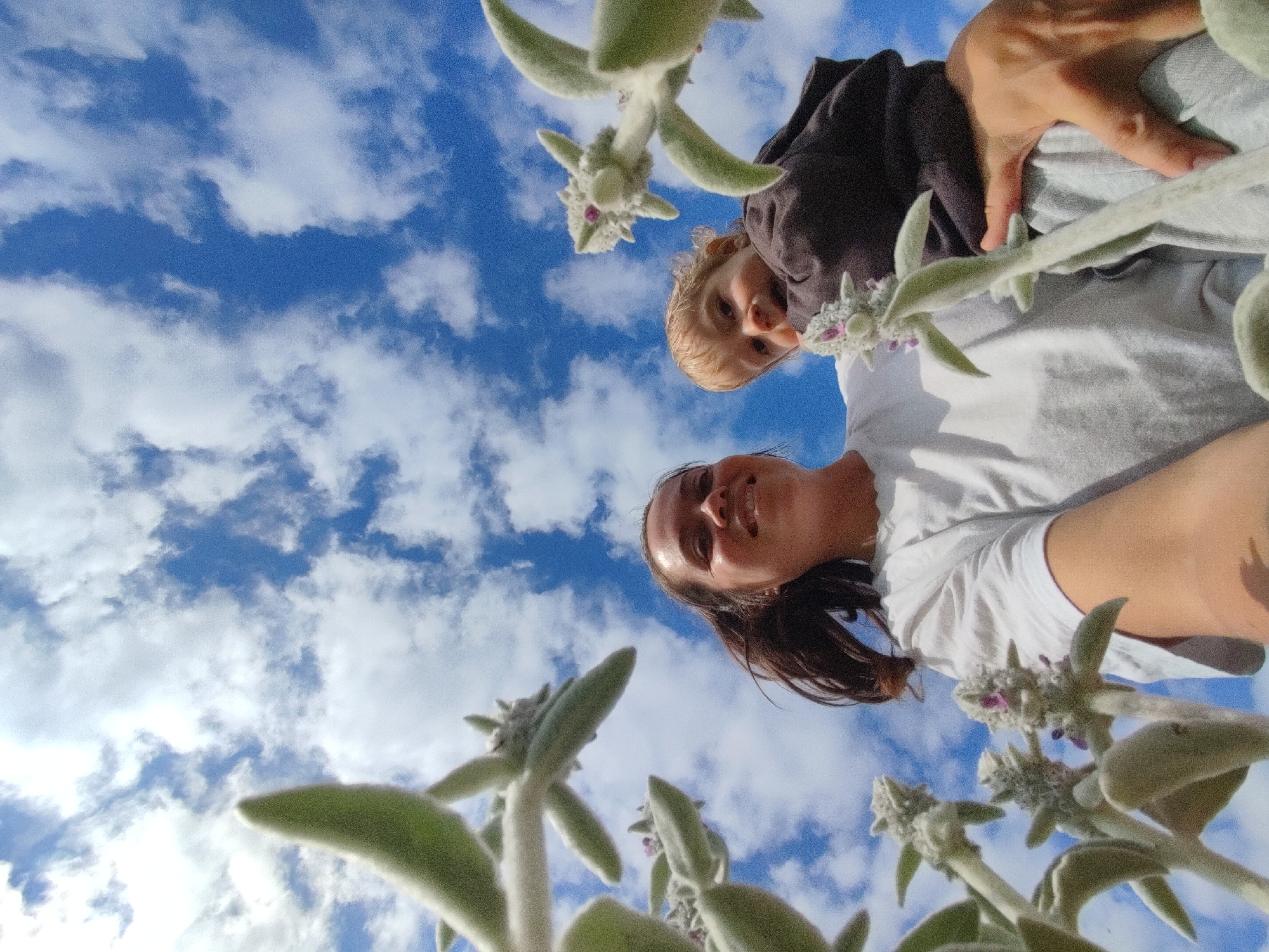 In an outdoor scene with a blue sky and white clouds, a person with dark hair smiles upwards towards the camera. They are wearing a white t-shirt. A child with blonde hair is positioned above and to the left of the smiling person, also looking towards the camera. The child is wearing a dark hooded garment. The image is taken from a low angle, with fuzzy green plants in the foreground, obscuring parts of the sky and the subjects. The overall impression is a candid, close-up shot with the subjects looking up into the sky. There is no visible indication of the city of Фоки, Russia.