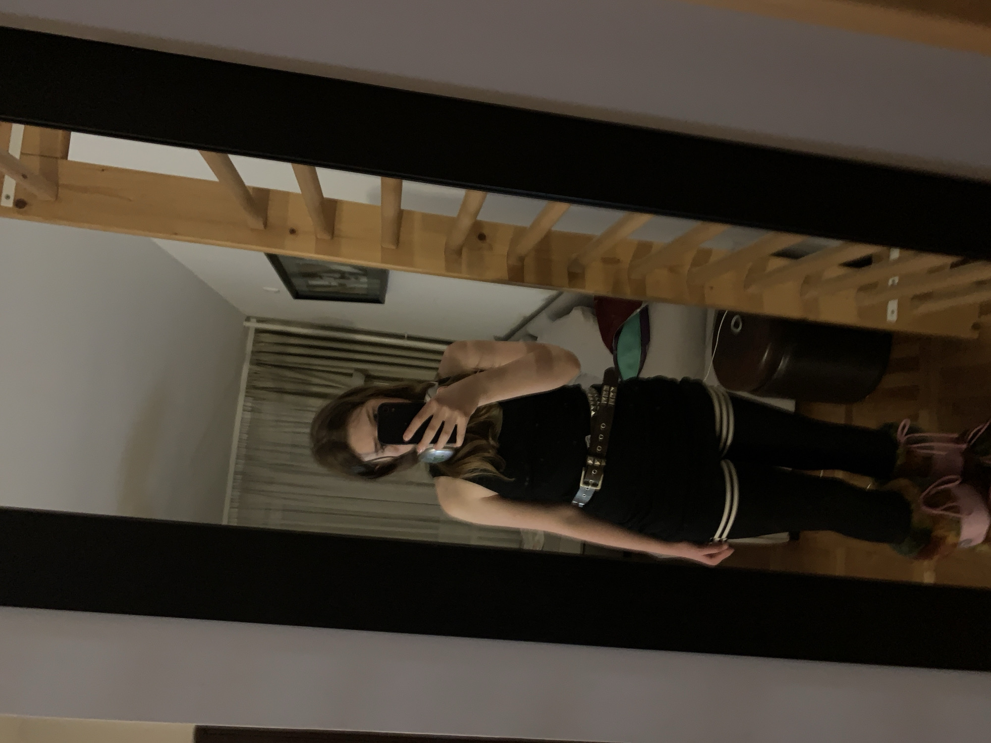 A person is taking a photo of themselves in a mirror. The person is wearing a black top, black pants with white stripes down the side, a studded belt, and pink shoes with fur. The room contains a wooden bunk bed, a framed picture, a white object, a curtained window, and a brown cylinder. The location is an interior space, likely a bedroom. The specific location in Vozdovac Urban Municipality, Serbia, cannot be determined from the image.