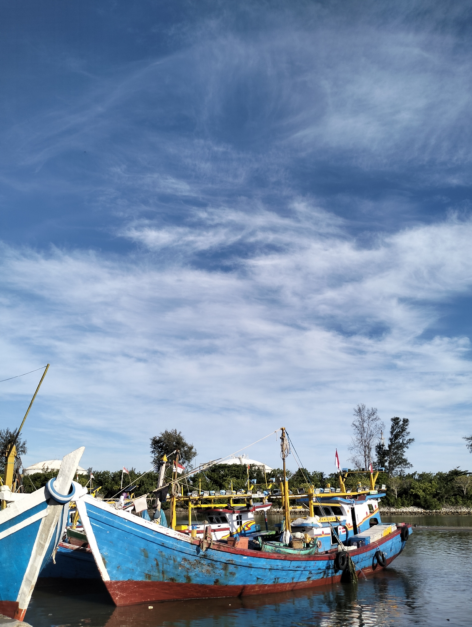 The image shows several blue and red wooden fishing boats docked in the river, with one boat prominently in the foreground. The boats have yellow accents and various rigging. The background features a blue sky filled with wispy white clouds. The riverbank is lined with green trees and shrubs. The location is Lhokseumawe, Indonesia.
