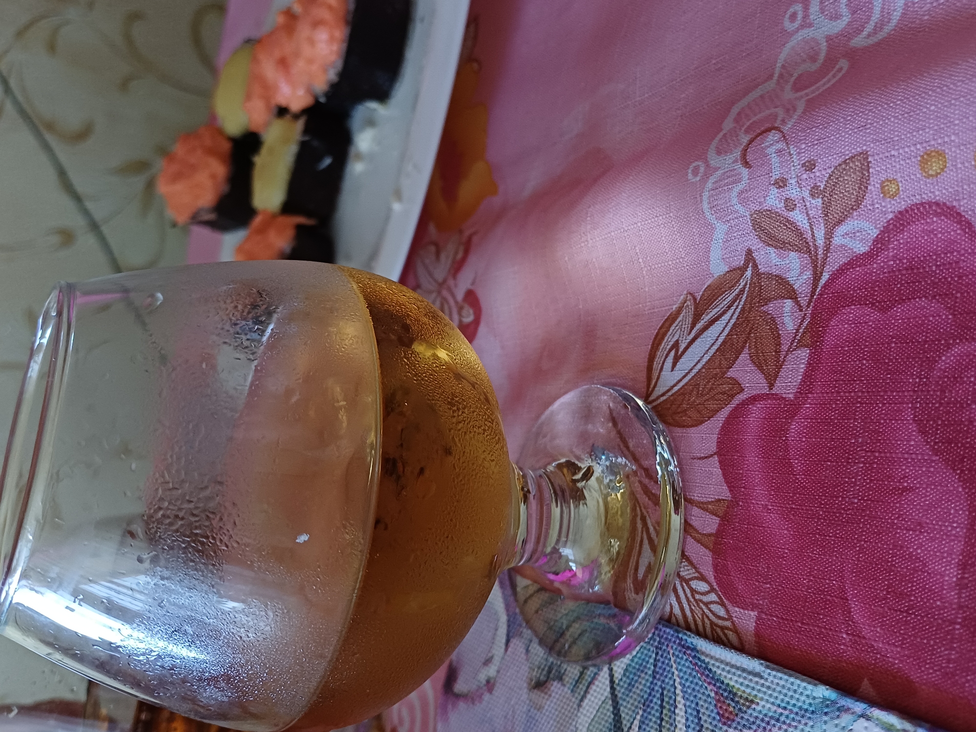 A brandy glass filled with amber liquid sits on a pink floral tablecloth. Condensation covers the exterior of the glass. In the background, a plate of sushi is visible. The overall setting appears to be indoors, likely a home environment, with a patterned wall in the distance. The lighting suggests it is daytime. No people are visible in the image.