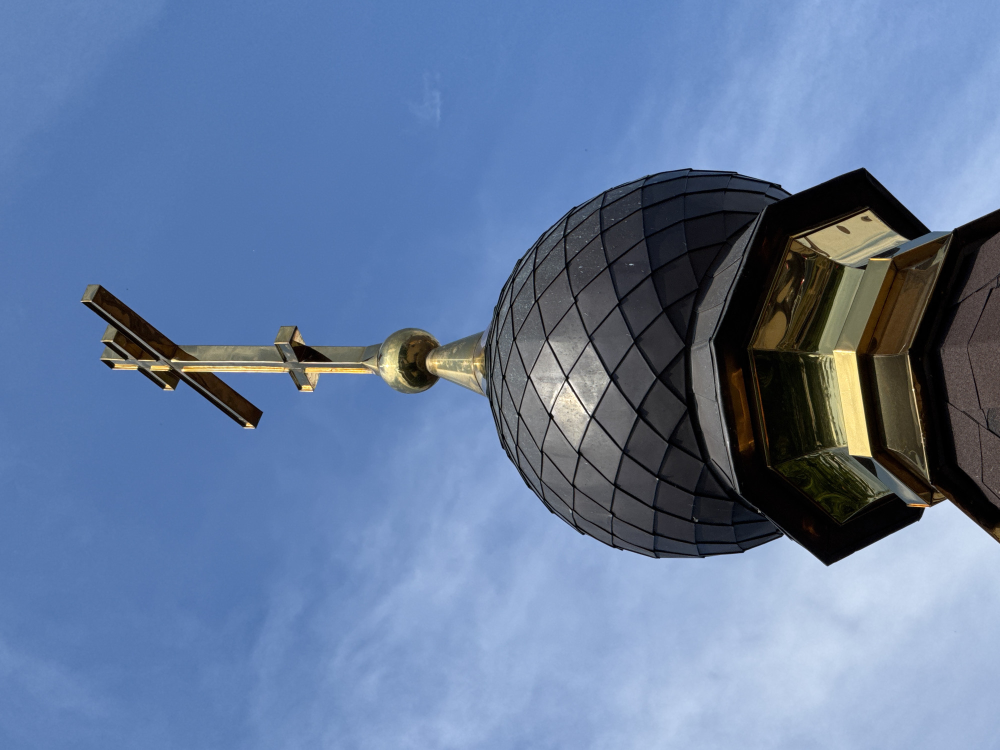 A close-up, low-angle shot captures the top of a church dome against a bright blue sky with wispy clouds. The dome is covered in dark, diamond-shaped tiles. A golden cross, positioned horizontally in the frame, stands prominently to the left of the dome. Below the cross and attached to the dome is a golden, bulbous finial. To the right of the dome, a section of gilded, angular architectural elements is visible, reflecting the surrounding environment. The clear sky suggests it is daytime, likely with fair weather. The image's perspective and the subject matter indicate it is a religious structure, possibly a Russian Orthodox church, given the context of Lomonosov, Russia. No people or text are visible.