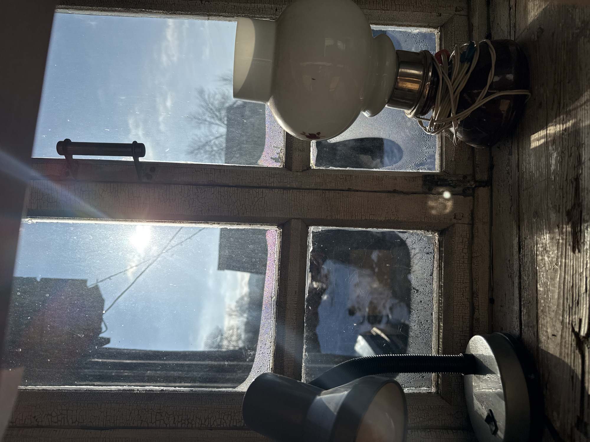 The image shows a weathered window with multiple panes, illuminated by sunlight. The window frame has peeling paint, revealing an aged surface. Through the glass, a blurry view of the outside is visible, including buildings, trees, and the sky. A white globe-shaped light fixture is mounted on the window frame, with electrical wires extending from it. A desk lamp is partially visible in the lower portion of the frame. There is no verifiable evidence the image was taken in Nizhnyaya Chermoda, Russia. There are no people visible in the image.