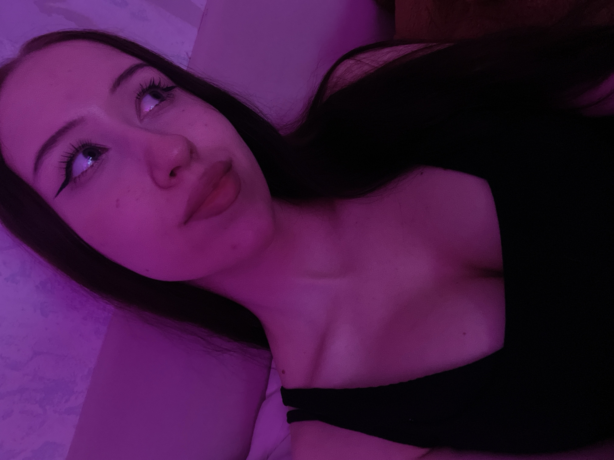 A young woman with long dark hair is shown in a close-up shot, illuminated by purple lighting. She is wearing a black top with thin straps. Her face is turned slightly to the right, with her eyes looking up and away from the camera. She has on winged eyeliner and visible eyelashes. The background appears to be a textured wall, also lit with purple light. The image does not contain any identifiable landmarks or details that would indicate a specific location in Votkinsk, Russia.