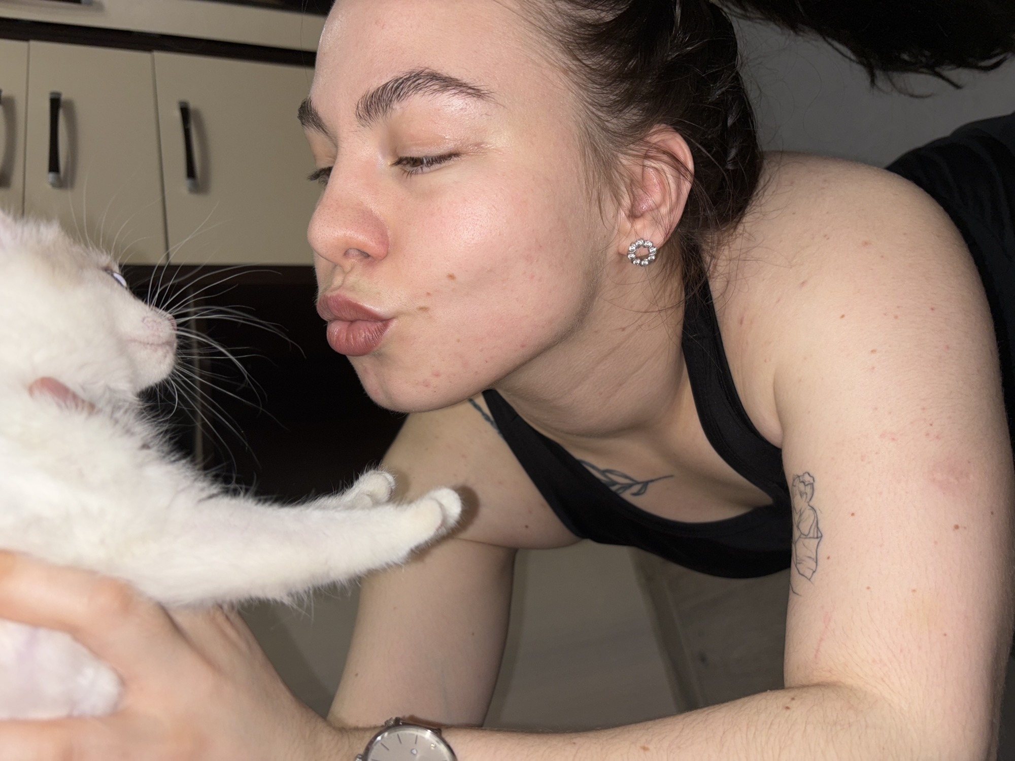 An indoor scene captures a tender moment between a young woman and a white kitten. The woman, appearing to be in her late teens or early twenties with dark hair pulled back, leans towards the kitten, her eyes closed and lips pursed in a kissing gesture. She wears a black tank top, revealing a small tattoo of a leafy branch on her chest and a larger outline tattoo of a classical figure on her right upper arm. A circular, clear-stoned earring is visible in her right ear, and a silver watch adorns her left wrist. Her skin shows some blemishes on her face and shoulders.
The small, fluffy white kitten, held gently by the woman's hand, has distinct whiskers and light blue eyes, with faint cream markings on its ears. The background features light-colored cabinets with dark handles, suggesting a domestic setting like a kitchen or utility room. The lighting is dim, indicating evening or nighttime. The image conveys affection and connection between the woman and her pet. No discernible text is visible within the image. The setting is contextualized as a residence in Perm, Russia.