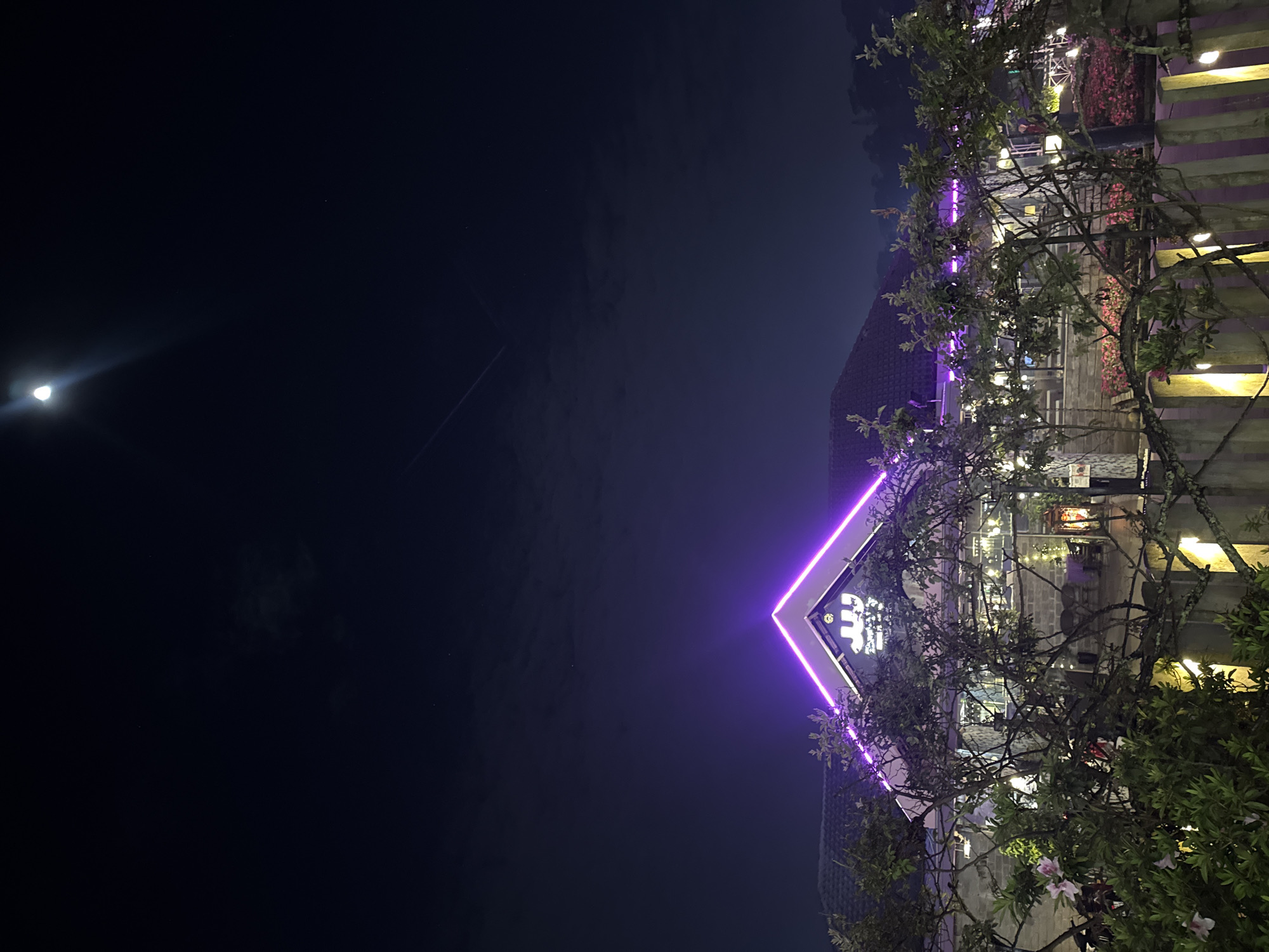 At night, the picture shows a building with bright lights in Da Lat, Vietnam. The moon is visible. A plane's contrail is in the sky. A triangular structure is outlined with purple neon lights. The building behind has windows with string lights. Vegetation is in the foreground.