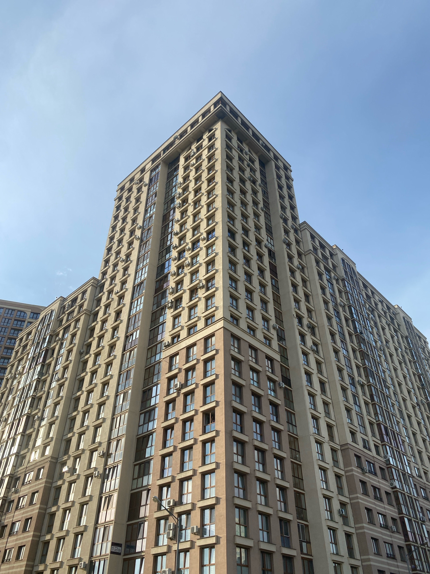 This is a low-angle, full-frame shot of a tall, modern apartment building in Minsk, Belarus. The building is constructed with light beige and brown facades, featuring numerous windows and balconies. Each floor is densely packed with windows, many of which are dark from interior lights or reflections. Several air conditioning units are visible mounted outside some of the windows. The building has a distinct, stepped architectural design, with a prominent cornice at the top. The sky above is a clear, pale blue, indicating daylight. To the left of the main building, another section of a similar apartment complex is visible, also showcasing rows of windows. The overall scene depicts a residential urban environment characterized by contemporary high-rise architecture.