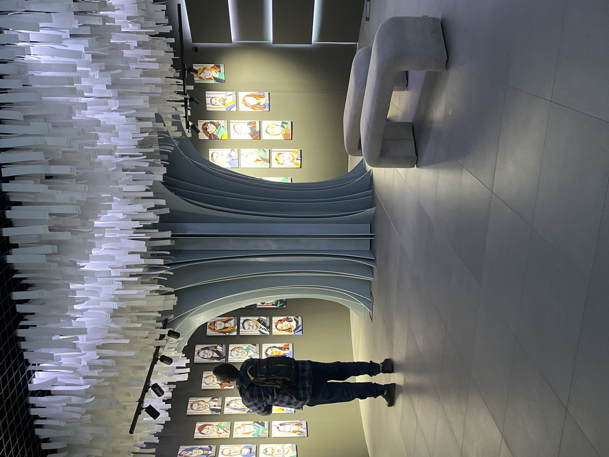 A man stands facing a wall adorned with portraits in an interior space. The wall is curved in an arch shape. Above the arch is a large installation of cascading white rectangular objects. A strip of lights are shining above the portrait wall, highlighting a modern gray couch. The interior space is located in the city of Moscow, Russia.