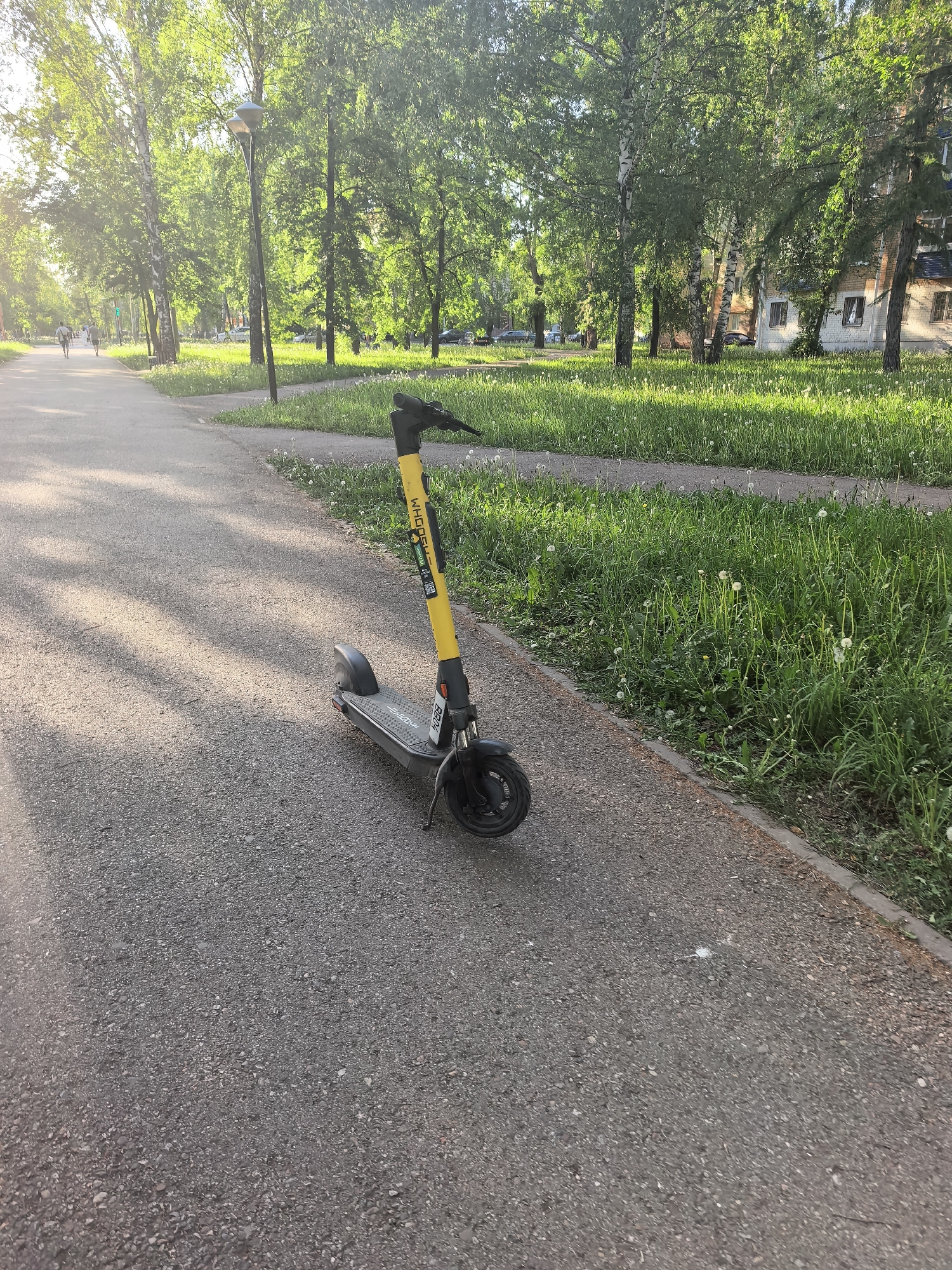 A yellow and black electric scooter stands on a paved path in a park. The scooter's side displays the word "WHOOSH". The path stretches into the distance, flanked by grassy areas and trees. A few people walk in the far distance on the path. A multi-story building is visible beyond the trees. The location is Sterlitamak, Russia.