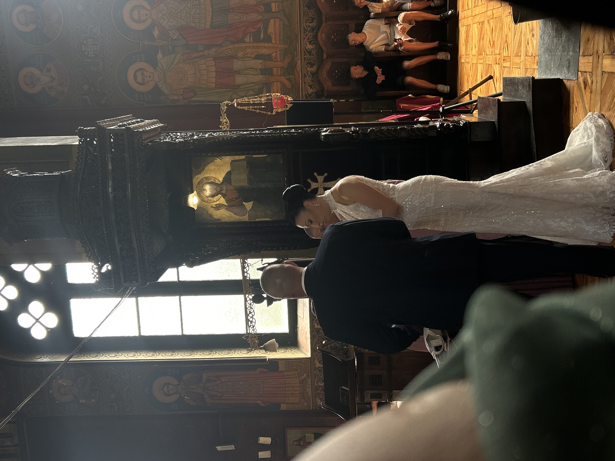 This image captures an indoor wedding ceremony taking place in an Orthodox Christian church, specifically identified as the **St. Nedelya Church in Sofia, Bulgaria**. In the foreground, slightly out of focus on the left, is a person's shoulder and back, wearing a green garment. The primary subjects are the bride and groom, positioned centrally, with their backs mostly to the viewer for the groom, and the bride's profile visible. The groom is a bald male, dressed in a dark suit (likely navy or black), standing with his back to the viewer and facing the bride. The bride is a female wearing a white, form-fitting, sleeveless mermaid-style wedding gown adorned with intricate lace or sequin detailing. Her dark hair is styled in an elegant updo, and she appears to be wearing earrings. She stands facing the groom, slightly to her left, with a serene and attentive expression. The church interior is richly decorated, characteristic of Orthodox architecture. The walls are extensively covered with frescoes (murals) depicting various saints. On the left wall, there is a circular icon of a bearded saint at the top, and a rectangular icon below it, clearly labeled in Cyrillic as "СТА ВАРВАРА" (Saint Barbara). On the right wall, two circular icons are visible at the top, one of which is labeled "СВ. КЛИМЕНТЪ" (Saint Kliment). Below these, two larger rectangular frescoes depict warrior saints, one in a green cloak and the other in a red cloak, standing upright. Intricate geometric and floral patterns are painted on the walls beneath the main frescoes. A tall, dark, and elaborately carved wooden structure, likely part of an iconostasis or a pulpit, stands between the main subjects and the right wall. This structure features an icon of Christ Pantocrator with a golden halo, holding a Gospel book. A small, ornate hanging metal vigil lamp with a red glass insert is suspended from a bracket attached to this wooden structure. Natural light streams into the church through tall, rectangular windows on the left side, which feature a distinctive quatrefoil (four-leaf clover) shape at their top. Bright light from these windows creates highlights and shadows within the scene. Thin wires are visible stretched across the window on the far left. On the far right, three young men are seated on a wooden bench or pew, casually dressed in t-shirts and shorts, observing the ceremony. The flooring throughout the church is a wooden