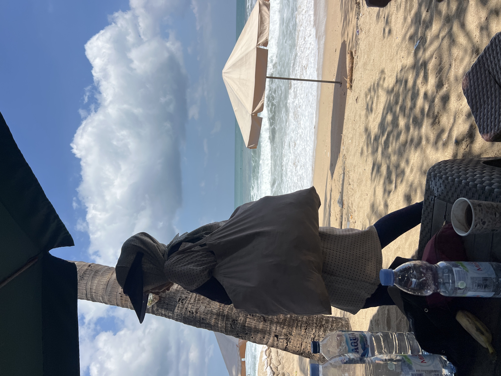 A medium shot captures a woman from behind walking on a sandy beach in Kuta, Indonesia, with the ocean and sky as the backdrop. She is wearing a light-colored patterned tunic, dark leggings, and a sun visor, with a large tote bag slung over her shoulder. To her left, a tall palm tree stands partially shaded by a dark green umbrella. In the background, a beige beach umbrella stands open on the sand, with waves gently rolling onto the shore under a bright blue sky dotted with white, puffy clouds. In the foreground, a woven table holds several plastic water bottles, a banana, a dark bag, and a paper cup. The scene is illuminated by natural sunlight, casting shadows from the umbrella and palm tree across the sand.