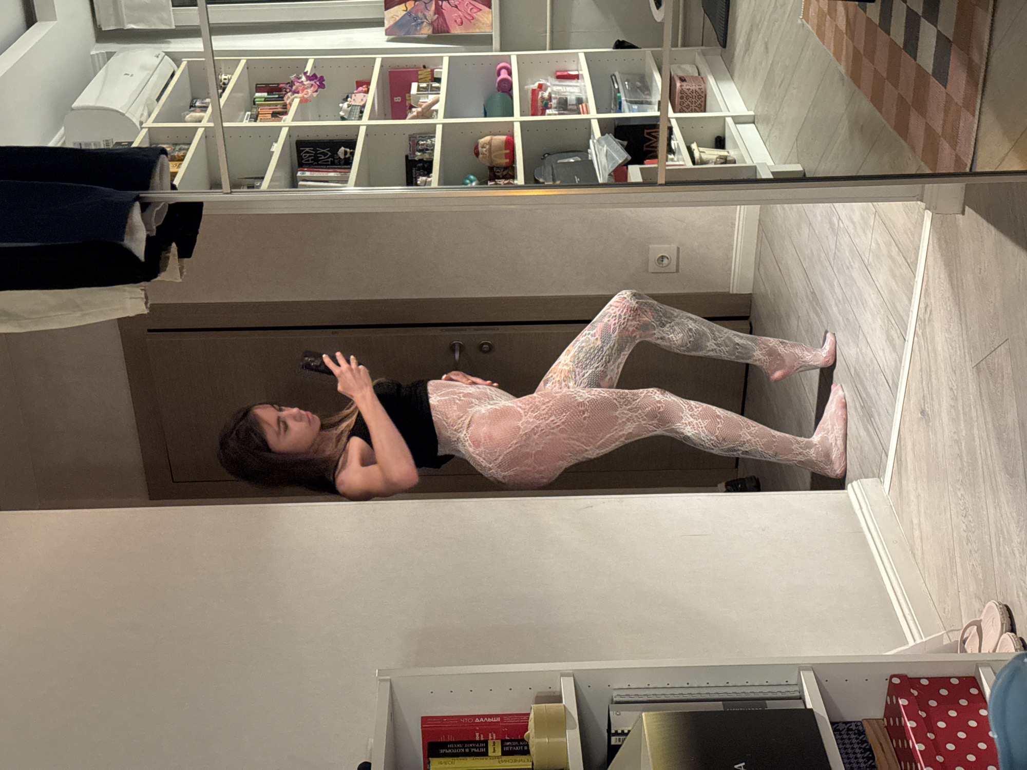 The image shows a young woman standing in what appears to be a room, possibly a dressing room or bedroom, with a large mirror reflecting part of the scene. The woman is wearing a black tank top and white lace-patterned tights. She is holding a smartphone in her right hand and appears to be taking a selfie. Her left hand is resting on her hip. The room has light-colored walls and wooden flooring. To the left of the woman, a white bookshelf is visible, filled with books and other items. To the right, reflected in the mirror, is another tall white shelf unit with various objects, including books, decorative items, and storage containers. An air conditioning unit is mounted on the wall above the shelves. A doorway is visible behind the woman. The overall lighting suggests an indoor setting, possibly at night or with dim natural light. The presence of the mirror and the woman's action of taking a selfie indicate a personal or private space.