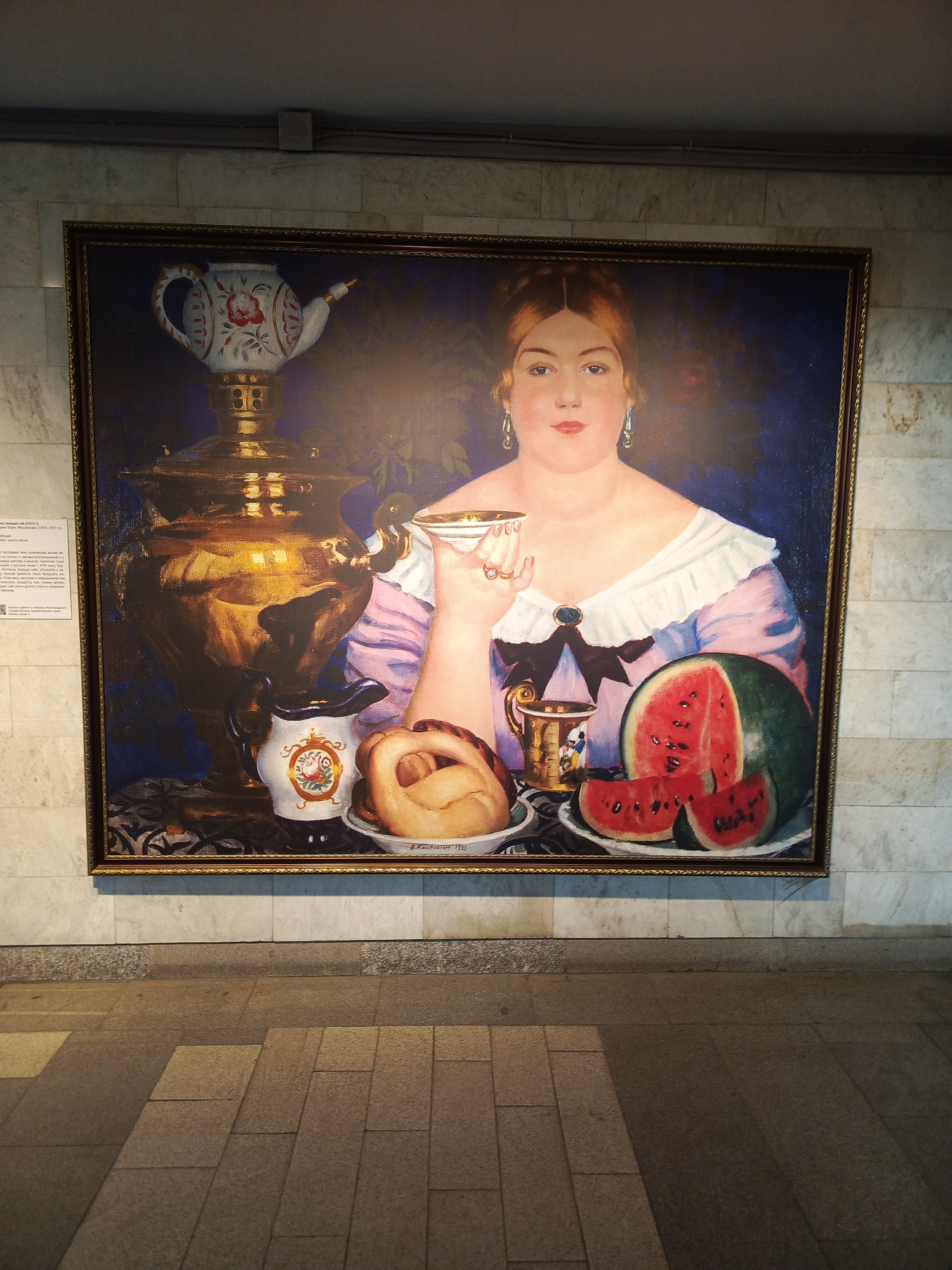The image shows a framed painting of a woman seated at a table with a samovar, teapot, teacup, plate of pastries, and watermelon. The woman is light-skinned with auburn hair and is wearing a light pink dress with a white lace collar. The background of the painting is dark blue. The painting is hanging on a light-colored marble wall with a horizontal stripe of darker stone near the bottom. The floor in front of the painting is paved with gray rectangular stones. The location is likely in Nizhny Novgorod, Russia, as paintings with similar subjects are often displayed in Russian museums.