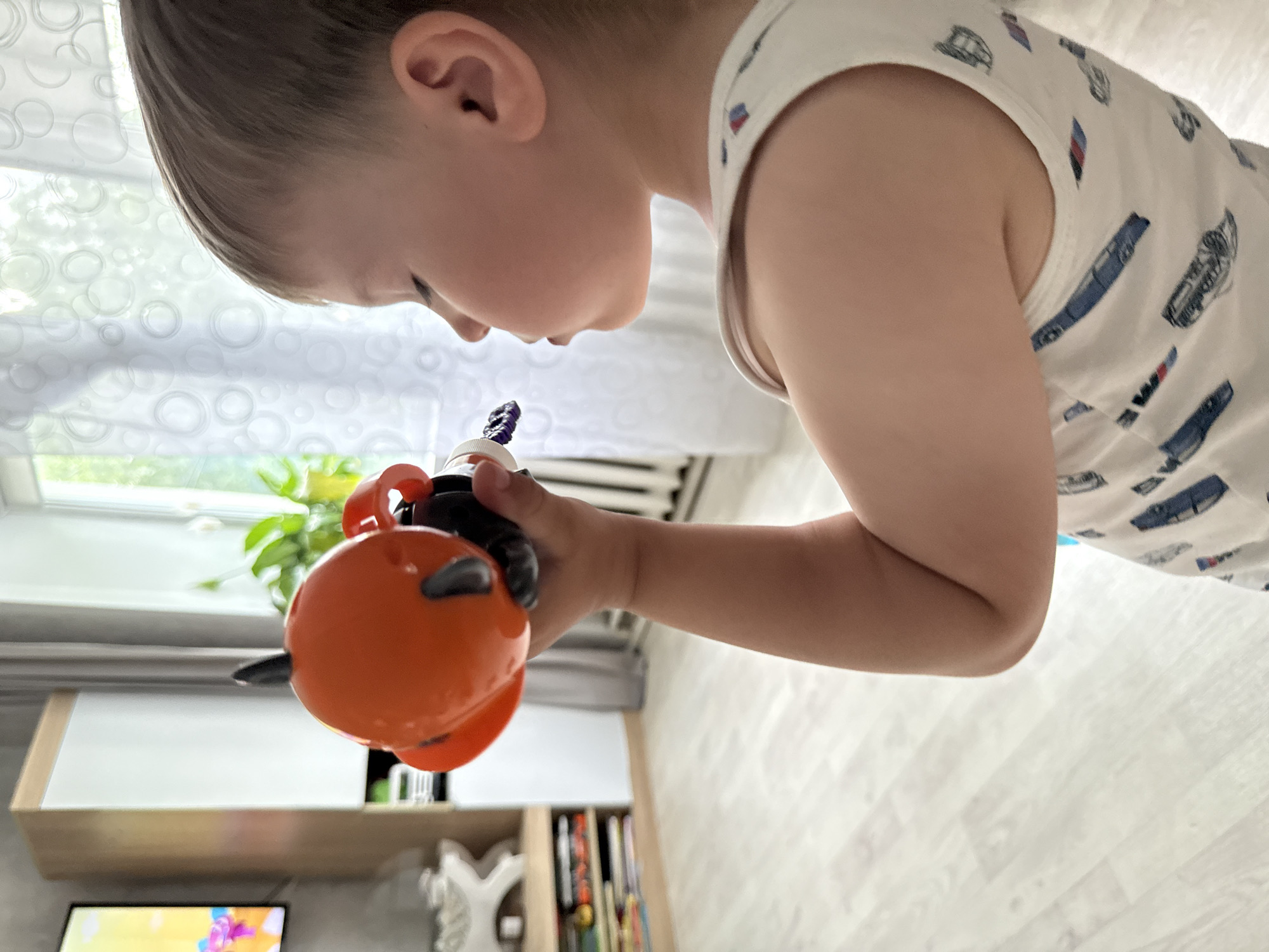 A young boy, with fair hair and closed eyes, is holding an orange toy with black accents near his face. He is wearing a white tank top with a pattern of blue cars and red and blue accents. The boy's left arm is bent and raised, holding the toy. The background shows a room with light-colored wooden floors, a white cabinet with a wooden top, and a television screen displaying a colorful image. A window with white sheer curtains is visible in the background. The scene appears to be indoors, likely a living room or play area. There is no specific information about the location in the city of Октябрьское, Russia.