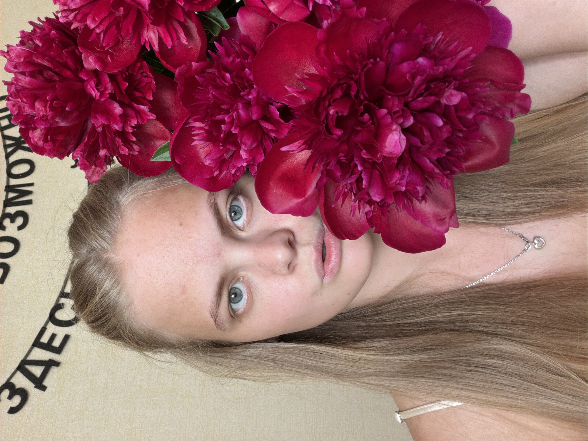 The image is a close-up, vertically oriented portrait of a young woman surrounded by a lush bouquet of deep red or magenta peonies. She has long, straight, light blonde or brown hair, fair skin, and striking blue eyes that look directly at the camera. Her lips are slightly parted, and her expression is calm and direct. Several large, full peony blossoms are positioned around her head and face, with some partially obscuring her hair and one touching her cheek. A delicate silver necklace with a small heart-shaped pendant is visible on her collarbone.
The setting appears to be indoors, against a light-colored background that could be a wall or a textile. Black Cyrillic text is visible on this background. Partially legible words include "ВОЗМОЖНО" (Vozmozhno, meaning "Possibly" or "Perhaps") and "ЗДЕСЬ" (Zdes', meaning "Here"). The image captures a posed moment, focusing on the woman's features and the vibrant flowers. The time of day is not discernible from the indoor lighting. Given the Cyrillic text, the location context provided, Izhevsk, Russia, is consistent with the visual cues.