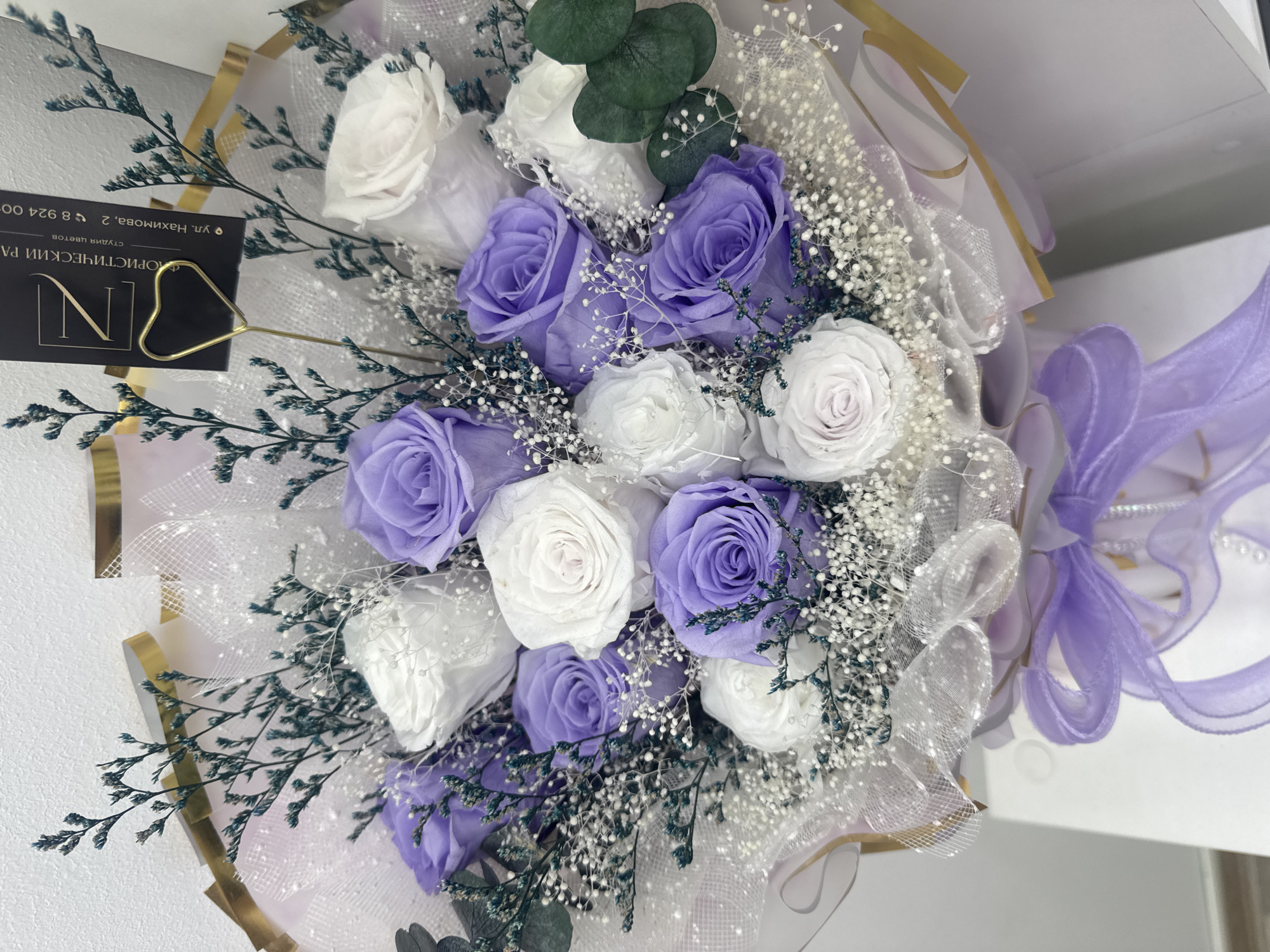 This is a medium shot, eye-level, close-up photograph of a bouquet of preserved roses, predominantly in white and lavender hues, arranged with dried floral accents and baby's breath. The bouquet is wrapped in translucent white mesh and possibly cellophane, with hints of purple and gold decorative paper peeking through. A lavender ribbon is tied in a bow at the bottom of the bouquet. A black business card with gold lettering is visible in the upper right portion of the bouquet, advertising "FLORISTIC ROOM" and a phone number. The background is a plain, light-colored wall. The photograph does not depict any discernible location within the city of Komsomolsk-on-Amur, Russia; it is a studio or indoor shot focusing solely on the floral arrangement. There are no people or animals present.