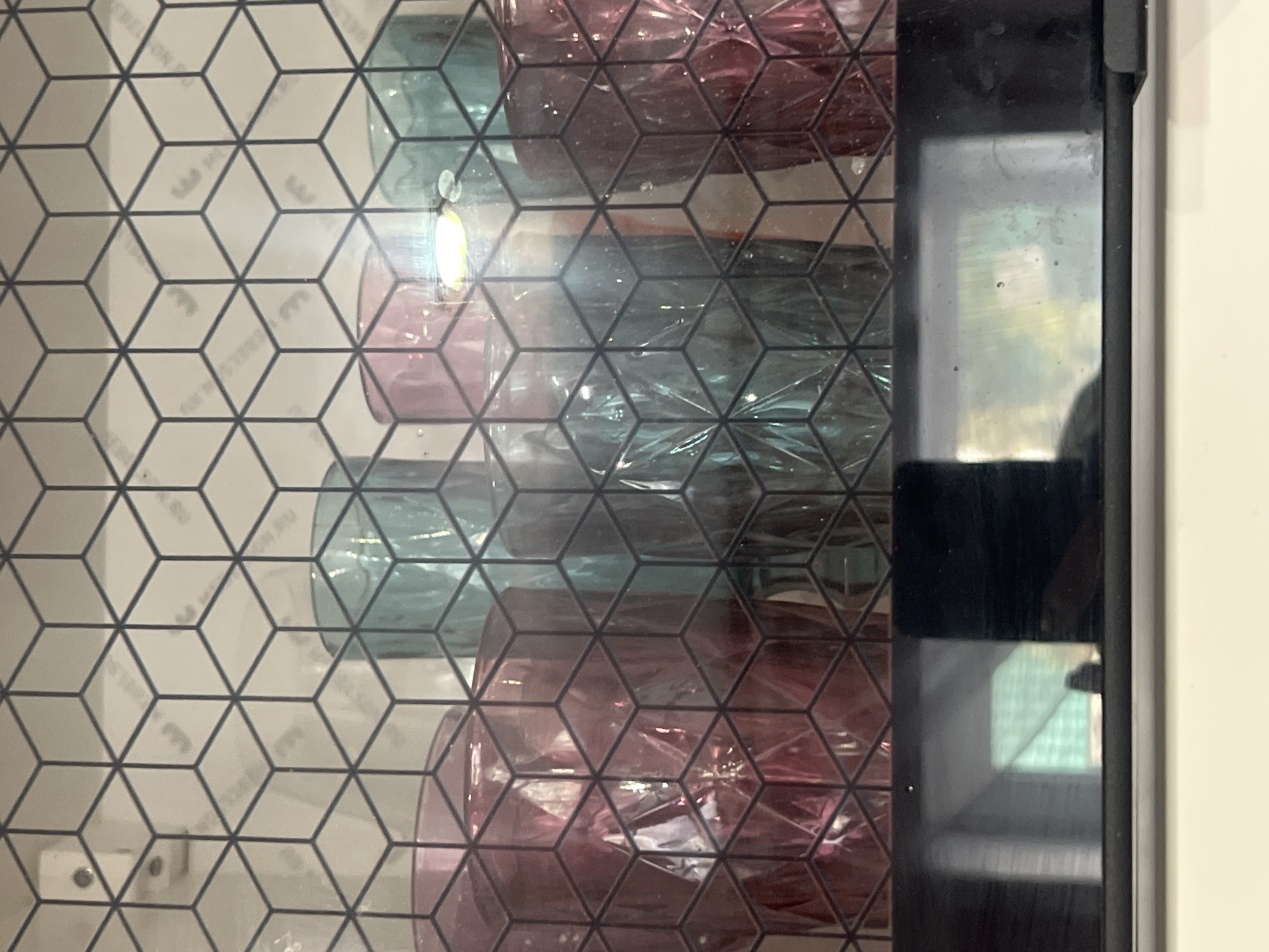 The image shows a close-up view through a glass door with a geometric pattern. Inside the cabinet, there are several decorative glass objects, possibly vases or bottles, in shades of pink and blue. The door has a black frame and reflects some external light sources. The location, Boguchar, Russia, cannot be confirmed or refuted based solely on the visual information in the provided image.