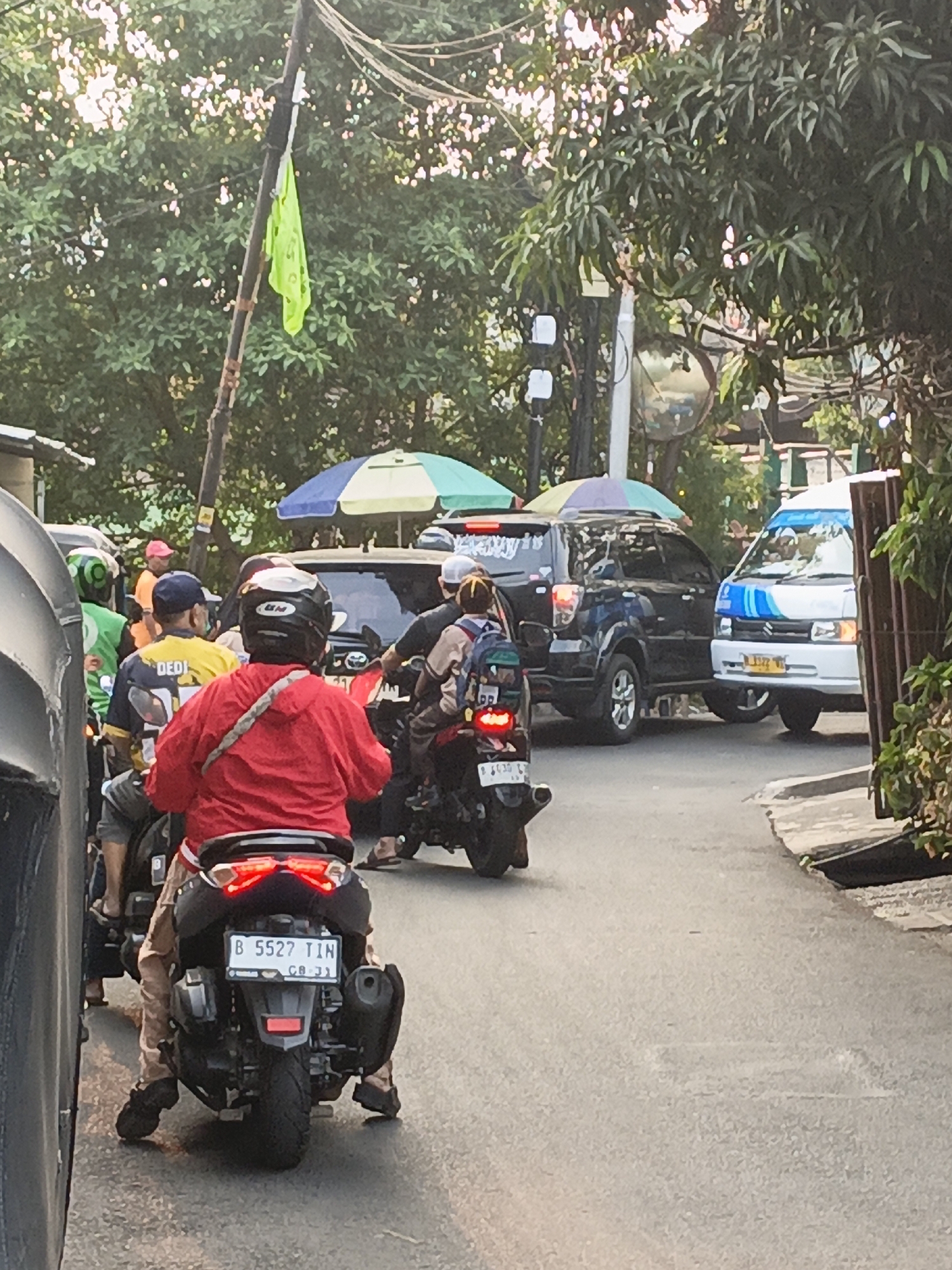 This is a medium-shot, eye-level, vertical-format photograph of a congested street scene in East Jakarta, Indonesia. The perspective is from behind a scooter rider wearing a red jacket and black helmet. The scooter's license plate reads "B 5527 TIN".
In the foreground, the back of the scooter and its rider are prominent. To the left, a portion of another vehicle, possibly a car, is visible. Further back, to the right of the scooter rider, another person is riding a scooter, wearing a backpack and a brown jacket.
In the middle ground, several vehicles are stalled in traffic. These include a dark-colored SUV, a white van with blue stripes and Indonesian license plates, and other motorcycles. There are also people visible on or near these vehicles, including a person wearing a bright yellow shirt with "DEDI" printed on it. Two colorful umbrellas, one blue and white, and another green and blue, are set up behind the SUV.
The background consists of lush green trees, partially obscuring overhead electrical wires and poles. A bright green flag is attached to a wooden pole on the left side of the image. A large, reflective orb, possibly a mirror or a decorative element, is visible in the upper middle-right section of the frame. The street is paved and appears to be a typical urban road with some drainage. The overall lighting suggests it is late afternoon or early evening.