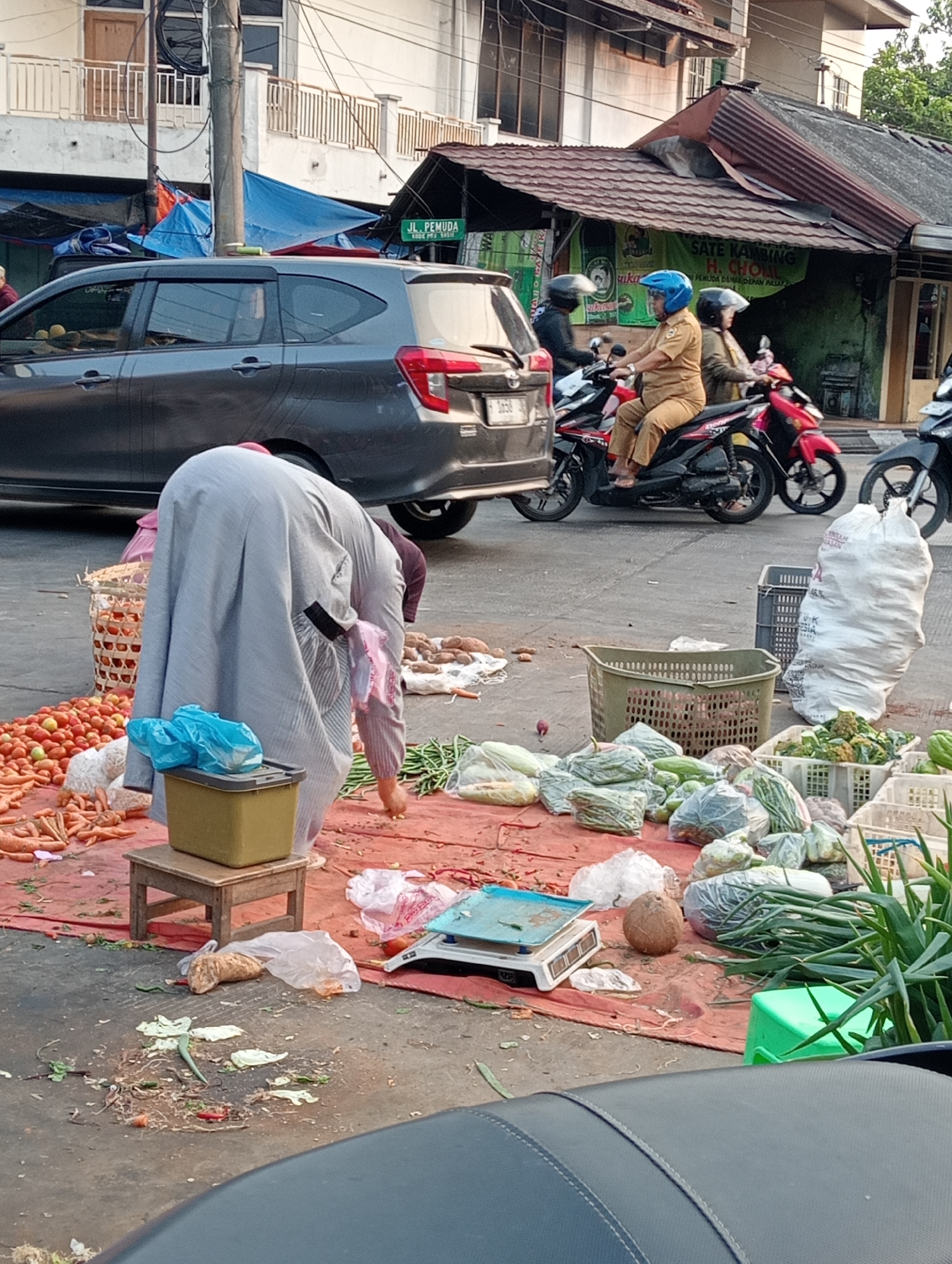 The image depicts an outdoor market scene in Demak, Indonesia, likely on a street or open area adjacent to buildings. A woman, dressed in a long gray garment covering her head and body, is bent over a collection of vegetables. She appears to be sorting or preparing produce, with her hands reaching towards a pile of green beans. In front of her is a small wooden stool with a yellow container on top, covered with a blue plastic bag.
The ground is covered with a reddish-brown tarp, on which various vegetables are displayed, including carrots, tomatoes, and leafy greens like cabbage and broccoli. Several woven baskets and plastic crates are used to hold some of the produce. A digital scale sits on the tarp near the center of the image. A brown coconut is also visible among the vegetables. Debris, including vegetable scraps and plastic bags, is scattered on the ground.
In the background, a dark gray car is parked, and several motorcycles are present. Two individuals wearing helmets are seated on motorcycles. One is wearing a light brown uniform. A street sign with the text "JL. PEMUDA" is visible, indicating the location. The overall environment suggests a busy marketplace with vendors and shoppers, typical of street-side commerce in Indonesian cities. The light suggests it is daytime, possibly late afternoon due to the shadows.