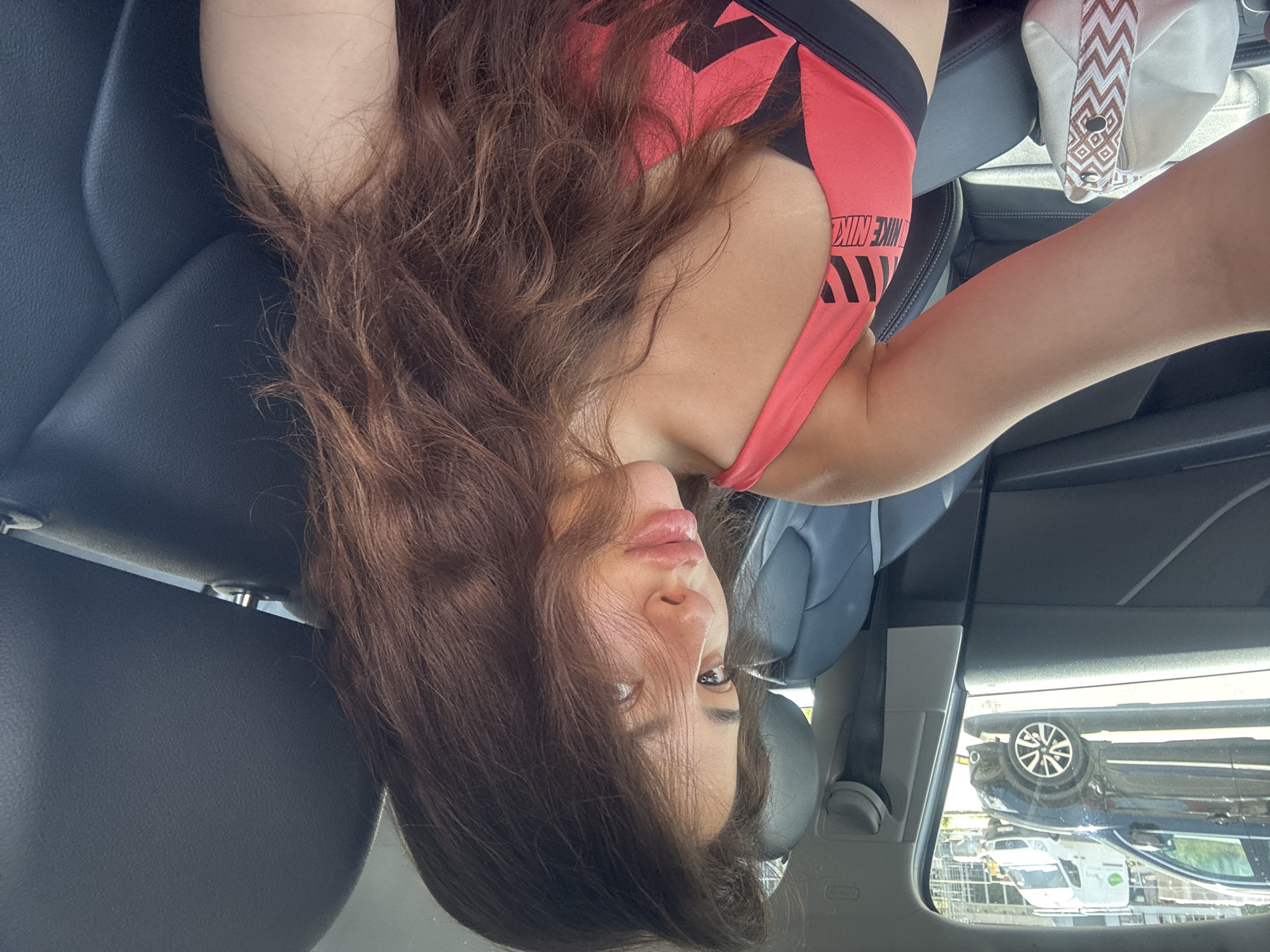 A young woman with long, brown, wavy hair is seated in the driver's seat of a car, facing the camera. She is wearing a pink sports bra with black lettering. Her left arm is resting on a white bag with a decorative patterned strap. The car's interior, with black leather seats and dark grey trim, is visible. Through the windshield, a black car and a white van are visible on a street, suggesting an urban environment. The background shows buildings under construction. The image appears to be a selfie taken in daylight. The specific location in Plovdiv, Bulgaria, cannot be determined from the visual information provided.