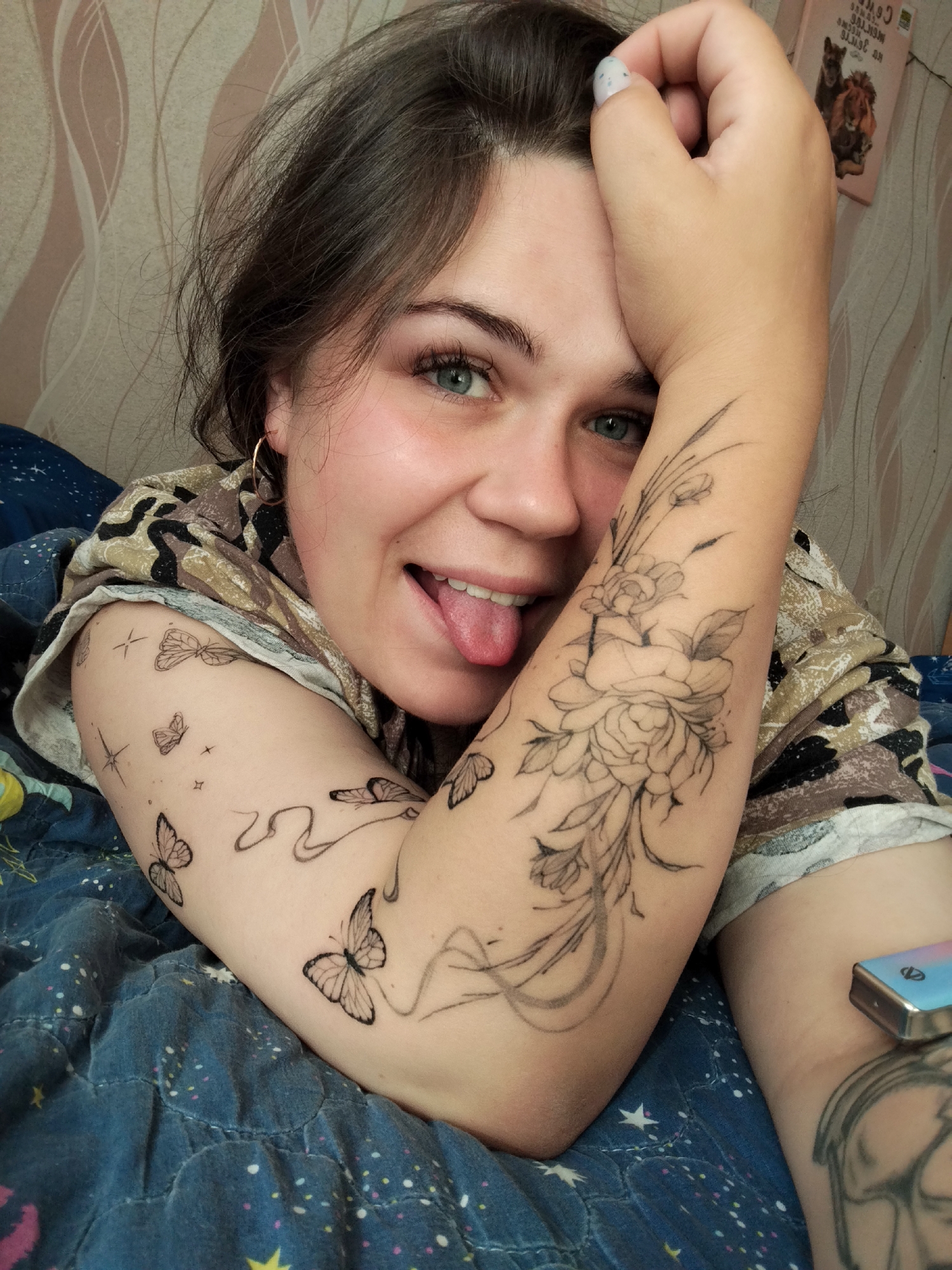The media file features a close-up shot of a young woman lying down, propped up on her arm. She is looking directly at the camera with a playful expression, her tongue sticking out slightly. Her eyes are a striking blue-green, and her dark hair is casually styled. The woman has several tattoos on her arm, including a detailed floral design and multiple butterflies. She is wearing a patterned shirt.
The setting appears to be indoors, possibly a bedroom, indicated by the presence of bedding with a celestial pattern and a wall with textured wallpaper. A small, rectangular object with a blue, pink, and silver casing is visible on her leg. In the background, a small pink book with images of animals is on a surface.
The overall mood is lighthearted and informal, suggesting a candid moment captured at home. There is no indication of the time of day or weather conditions. The location context is Chaikovsky, Russia, but there are no specific visual cues within the image to confirm this. No visible text is discernible in the image.