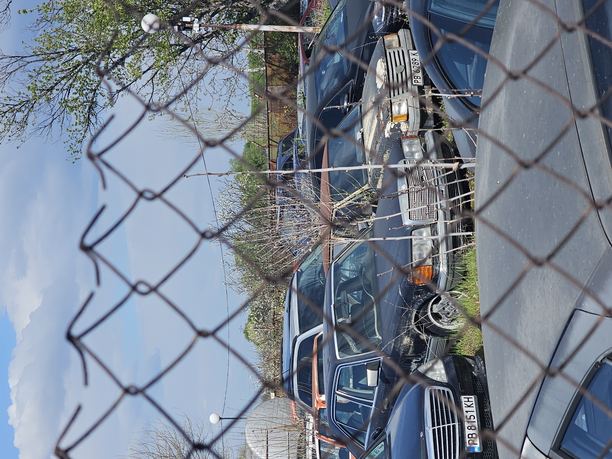 The image shows a view through a chain-link fence of several parked, apparently abandoned, Mercedes-Benz automobiles in varying states of disrepair. The cars are in a line, with some parked very close to one another. License plates are visible on some of the vehicles; one reads "PB 8151 KH," and another partially visible plate reads "PB 6289 K."
In the background, there are trees, a cloudy blue sky, and a street lamp. The location is suggested to be in Plovdiv, Bulgaria.