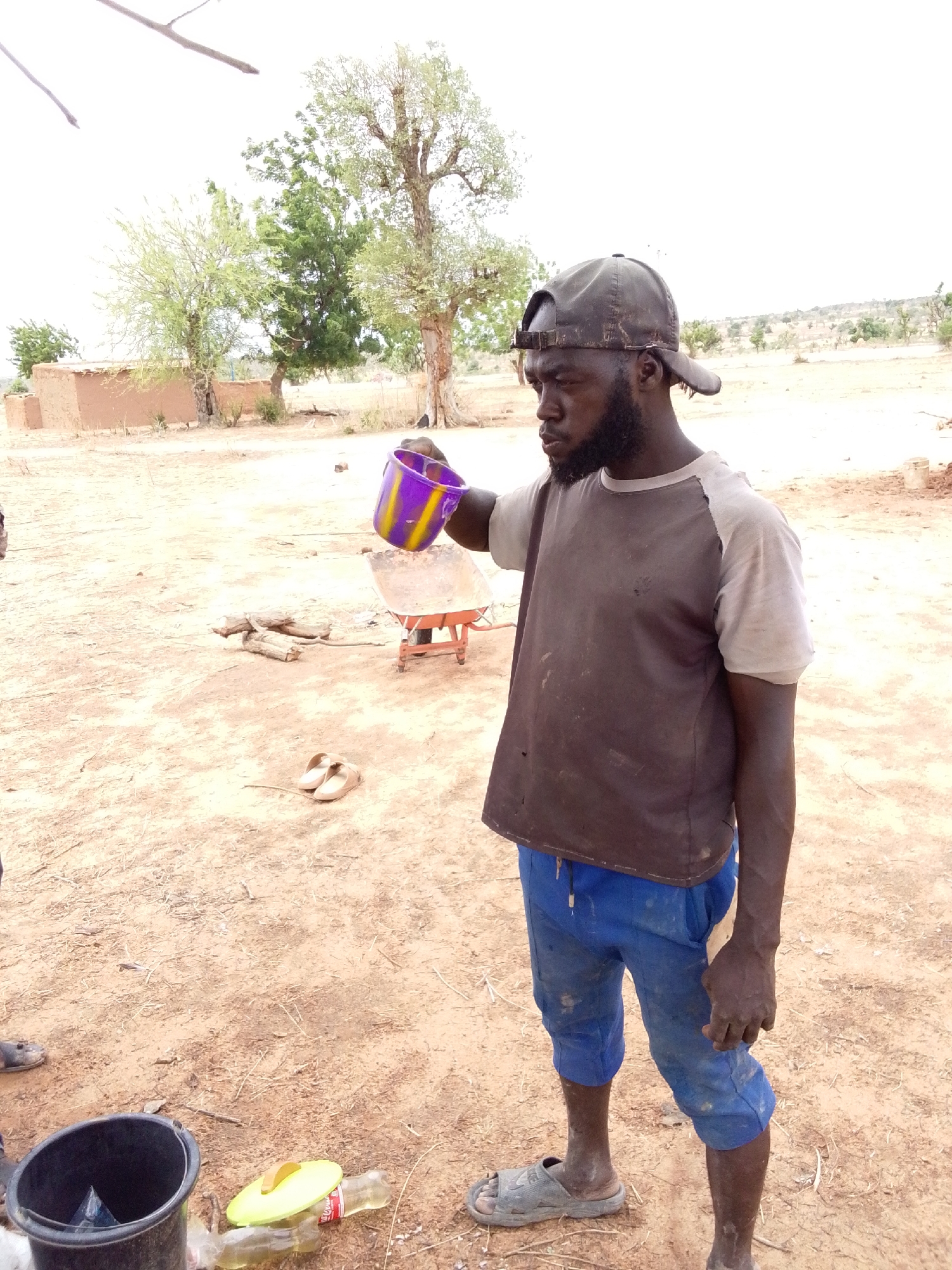The image depicts a man in Mulke Barikin Sayejo, Nigeria, standing outdoors in a dry, dirt-covered area. He is dark-skinned, bearded, and wearing a dirty baseball cap, a two-tone t-shirt (dark brown torso, light gray sleeves), blue cropped pants, and sandals. He is holding a purple and yellow plastic cup, tilting it as if pouring liquid. Behind him is a small red wheelbarrow and some chopped firewood. In the background, there are sparse trees, a small mud structure, and dry terrain. On the ground near the man's feet is a black bucket, a plastic bottle, and discarded items. The overall scene appears arid and rural.