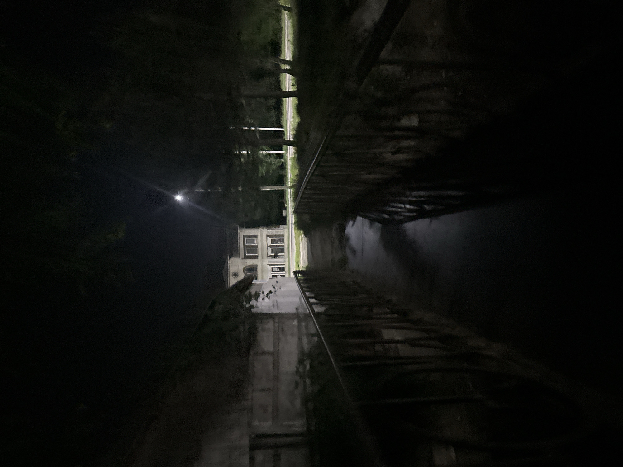 This image depicts a nighttime scene in Strășeni, Moldova. In the foreground, a narrow, slightly curved pedestrian bridge with dark metal railings spans a dark, reflective surface of water, possibly a canal or stream. The water reflects the ambient light from a distant streetlamp and the illuminated windows of a building.
The bridge leads towards a two-story building with a light-colored facade and arched windows on the upper level. This building appears to be a house or a small institutional structure, illuminated from within and by external light sources. To the left of the bridge and in front of the building, there's a rough, textured wall, possibly part of another structure, with some greenery growing on it.
Beyond the building and to the right, a road is visible, with trees lining its edge. A streetlamp stands to the left of the road, casting a bright light. The overall environment is dark, with foliage from trees partially obscuring the sky and creating shadows. No people are visible in the image. The scene suggests a quiet, possibly semi-rural or residential area within the city of Strășeni.