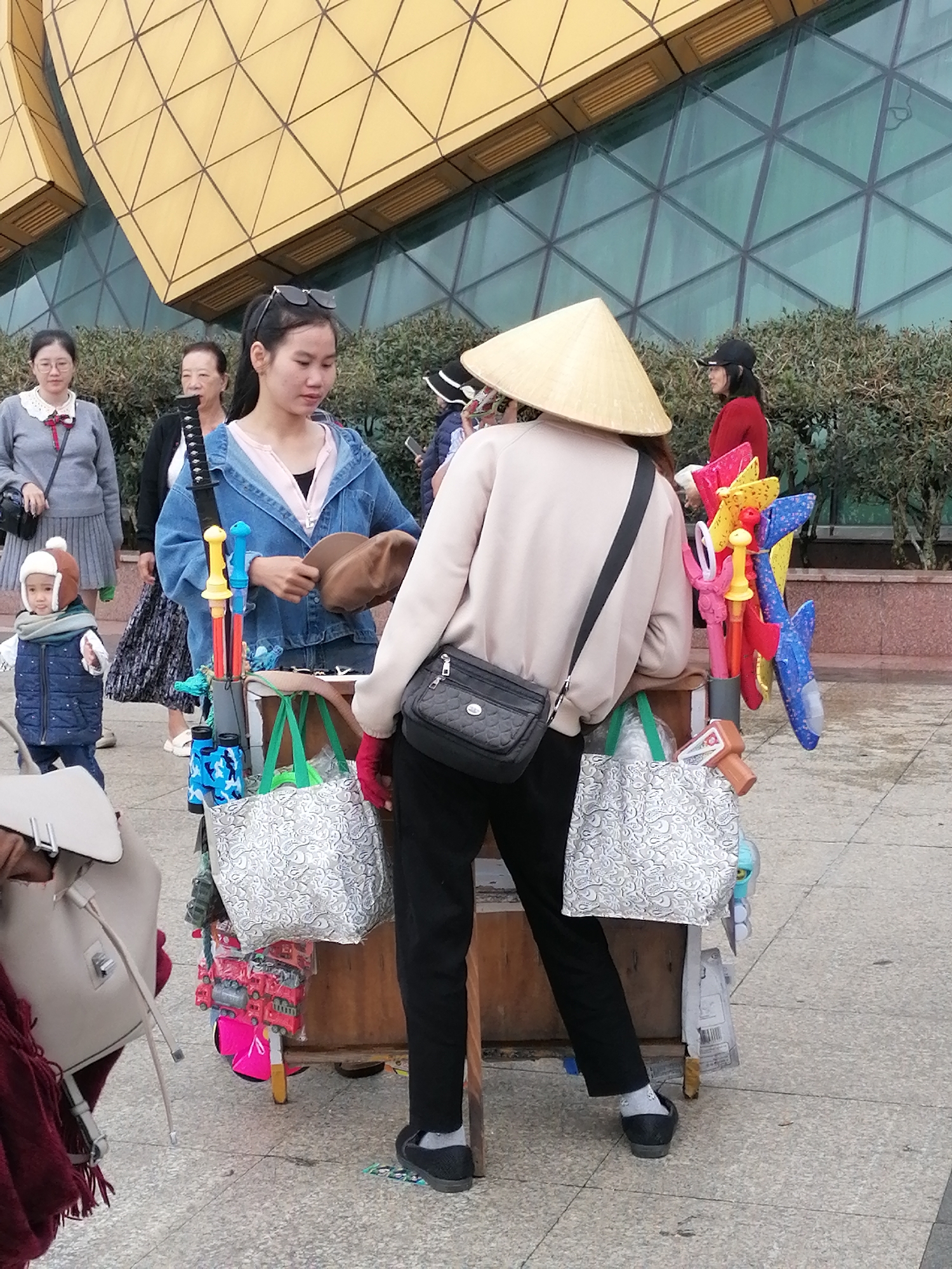 In Da Lat, Vietnam, two women, one wearing a conical hat, interact with a cart displaying colorful toys. The woman with the hat, dressed in a light-colored sweatshirt and black pants, faces away from the camera. She holds a brown hat. Her companion, in a denim jacket and a pink shirt, stands to her left. A young child in a blue vest and brown hat stands to the left of the cart. The cart is laden with various toys, including pinwheels, toy trucks, and other assorted items, all brightly colored. Behind them, the backdrop features a modern building with a distinctive golden, geometric facade and large glass windows. Lush green bushes line the area, indicating an outdoor setting. The ground is paved with light-colored tiles. The overall scene captures a bustling street-side transaction or interaction.