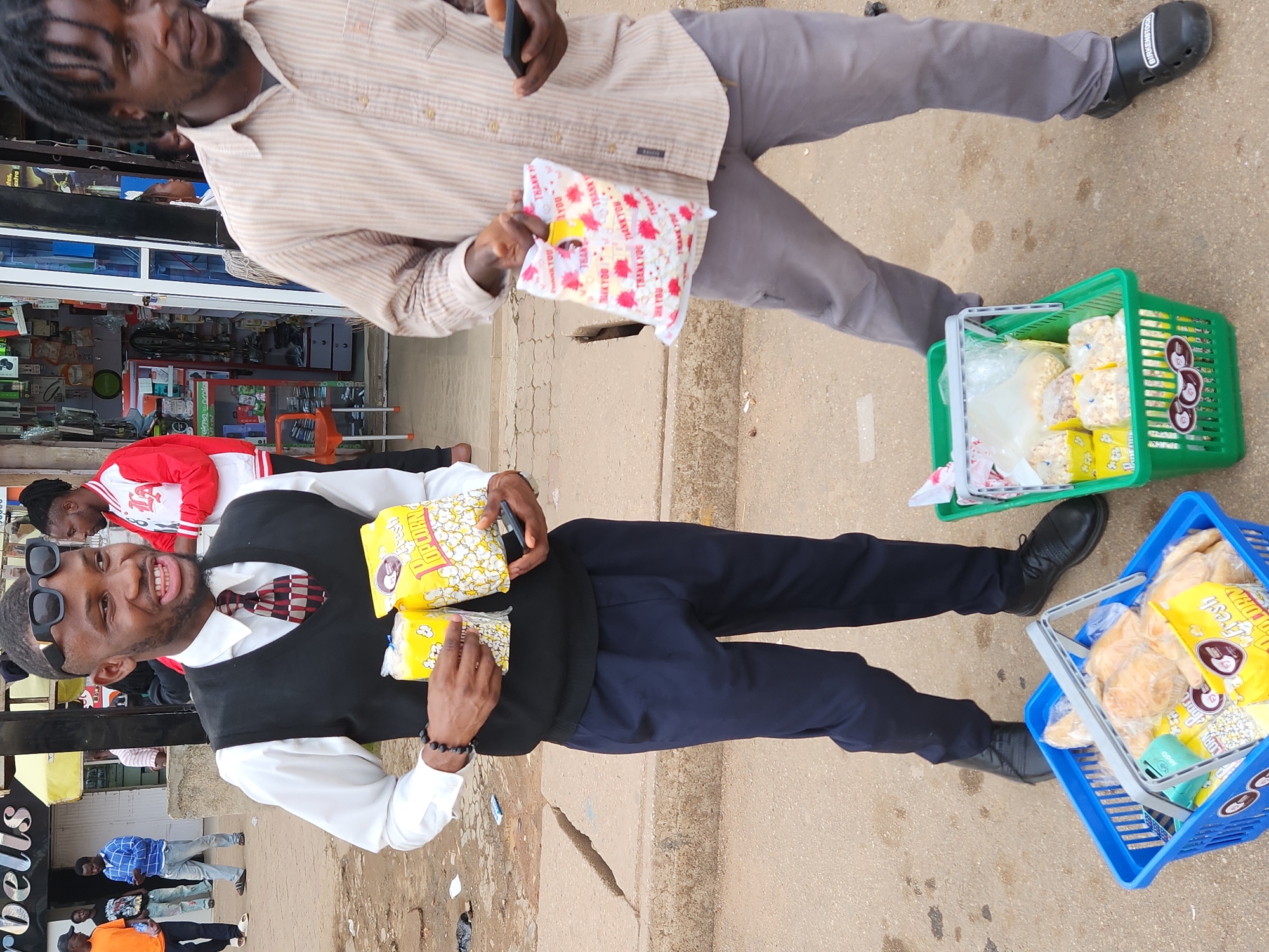 In the city of Jos, Nigeria, a young Black man with short dreadlocks and a beard stands in the foreground, wearing a light brown striped button-down shirt and grey pants. He is holding a small white plastic bag with red floral designs and the words "THANK YOU" printed on it in red. The bag appears to contain snacks. In his right hand, he holds a black smartphone.
To his left, another young Black man, dressed in a white shirt, black vest, and dark blue trousers, smiles and looks to his right. He holds two yellow boxes of popcorn in his hands. He is also wearing dark sunglasses perched on his head and beaded bracelets on his left wrist.
In front of the man in the striped shirt, a green plastic basket holds various bagged snacks, including popcorn. To the left of this basket, a blue plastic basket contains what appear to be bread rolls and more bagged snacks, including a yellow box of popcorn.
The background shows a street scene with a building that appears to be a shop. Several other individuals are visible in the background, including a man in an orange shirt, a man in a blue plaid shirt, and another man wearing a red and white jacket. The ground is paved with asphalt, and concrete curbs are visible. The lighting suggests it is daytime.