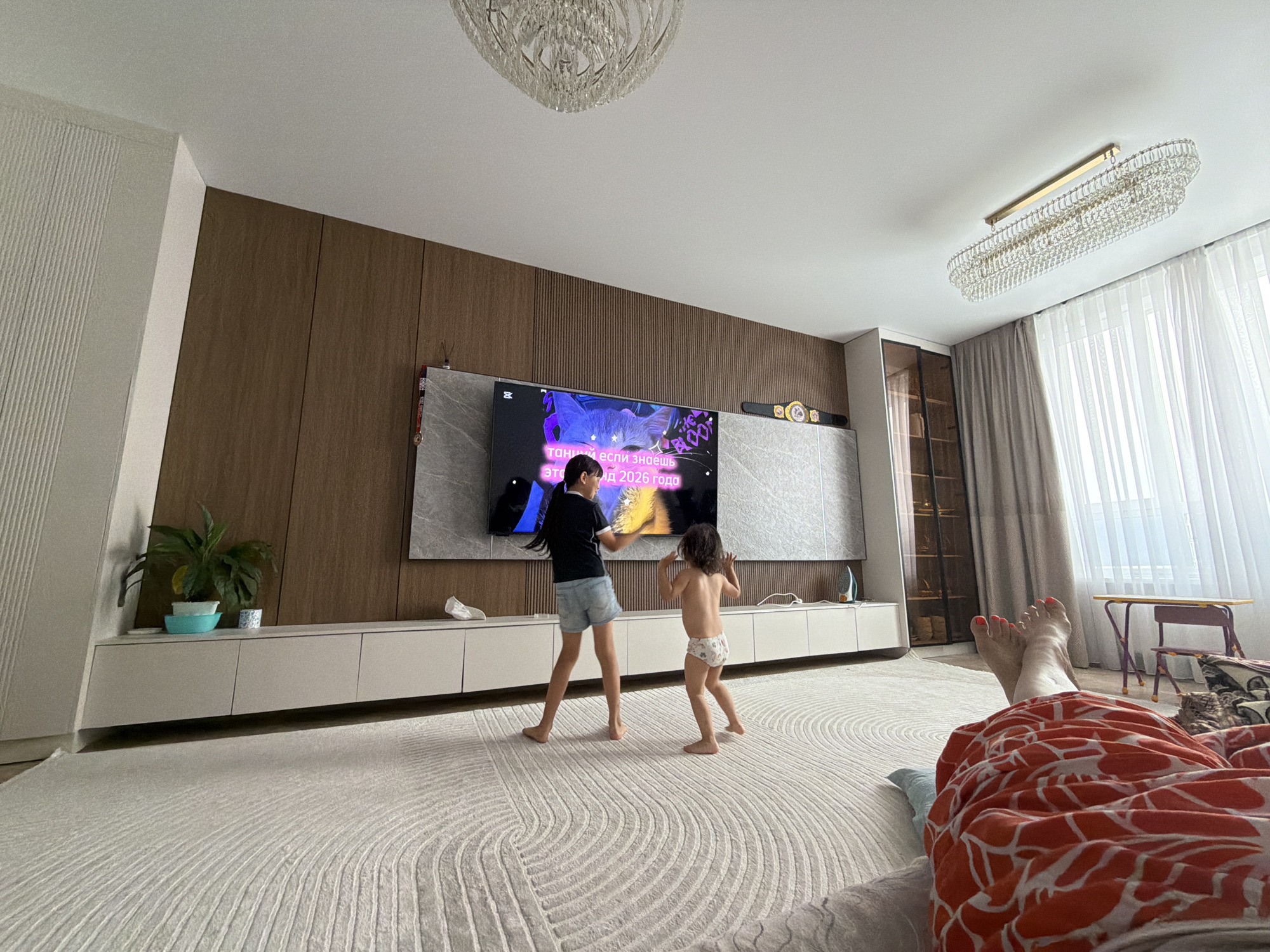 The image depicts a modern living room in Astana, Kazakhstan, bathed in natural light filtering through sheer curtains. Two young girls are dancing in front of a large television screen mounted on a wood-paneled wall. The older girl, wearing a black t-shirt and denim shorts, is on the left, while the younger girl, in a diaper, is on the right. The television displays text in Cyrillic script, which appears to be lyrics or a message related to dancing, possibly indicating a music video or a dance game. The room features a long, low-profile entertainment unit beneath the TV, with a potted plant on the left side. A plush, intricately patterned rug covers the floor. In the foreground, a person's legs and feet are visible, resting on a surface with an orange and white patterned fabric. The overall atmosphere suggests a casual, home setting, likely during the daytime.
