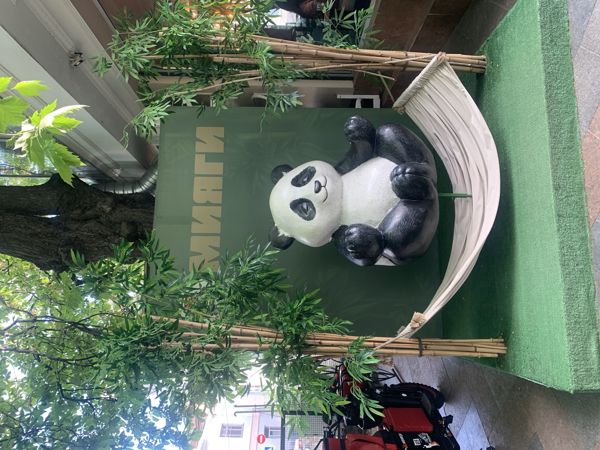 A vibrant outdoor commercial display features a large, black and white panda statue as its central element. The panda is depicted lying on its back in a white fabric hammock, with a relaxed and cheerful expression. The hammock is suspended within a setup that includes artificial bamboo stalks with green leaves, creating a mini-bamboo forest aesthetic. The background of the display is a solid green wall.
On this green wall, Cyrillic text is prominently displayed, which reads "ИМЯГГИ". The display rests on a green, carpet-like surface. To the left side of the scene, a large tree trunk and abundant natural green foliage are visible, suggesting an outdoor setting. Bright sunlight filters through the tree leaves, indicating it is daytime. The ground in front of the display is paved with patterned tiles.
In the lower right corner of the image, a red and black insulated delivery bag or box is present, resting on what appears to be a cart or scooter, possibly hinting at a food delivery service associated with the establishment. The overall scene is static, serving as an attractive and whimsical advertisement or decorative element for a business. No people are visible in the image.