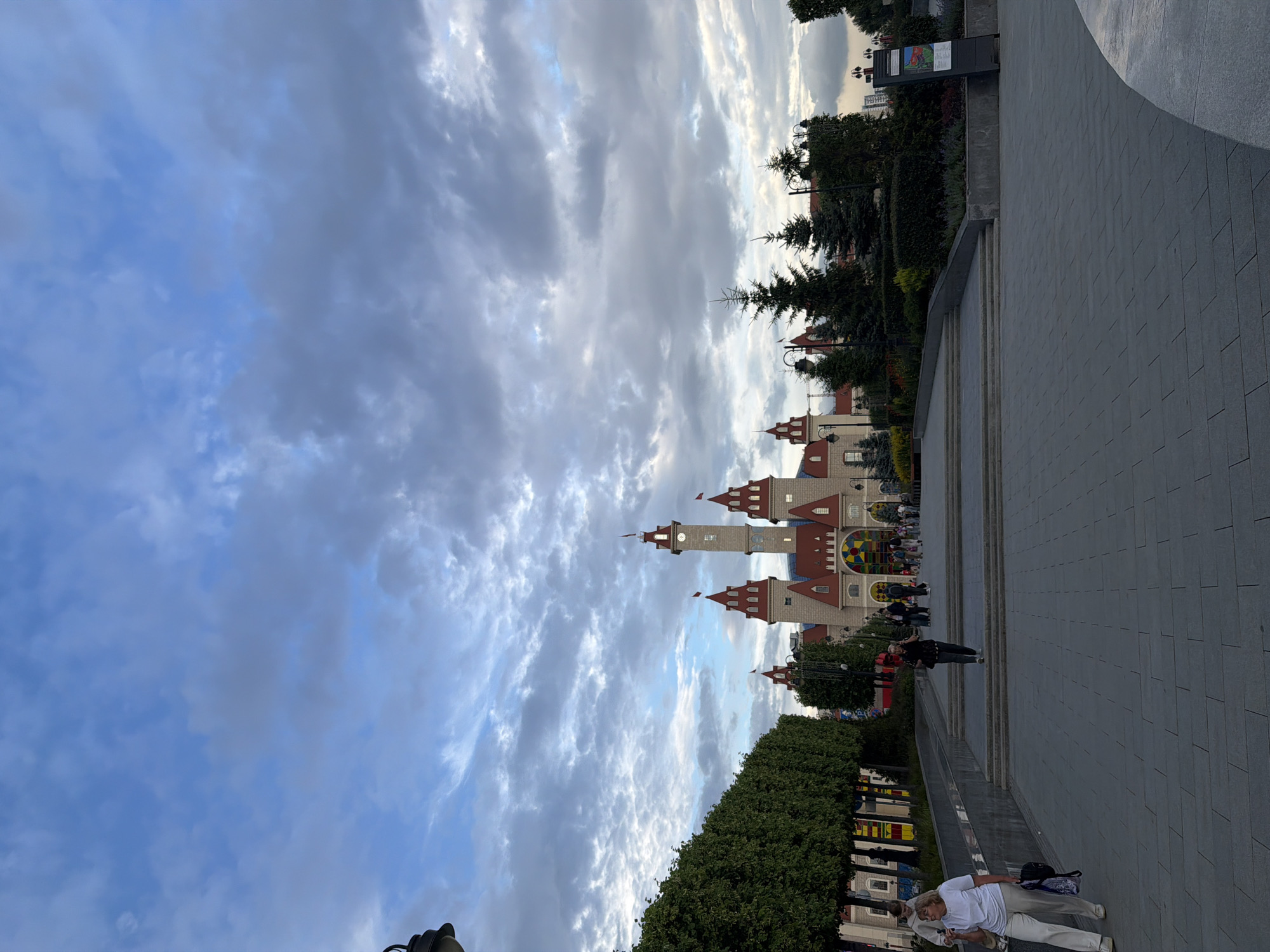 An exterior, eye-level, full shot shows an ornate, fairytale-like castle building with red-tiled roofs and towers under a cloudy, blue sky. The castle is situated in the background, with a wide, grey, paved plaza in the foreground. Several people are walking on the plaza. On the left, two women walk toward the camera, one in a white t-shirt and white pants, carrying a dark backpack, and the other in a black top and grey pants, talking on her phone. Behind them, a row of trimmed green trees lines the edge of the plaza. To the right of the castle, there are more trees and some architectural details of other buildings. The overall scene appears to be a public space, possibly an amusement park or a city square.