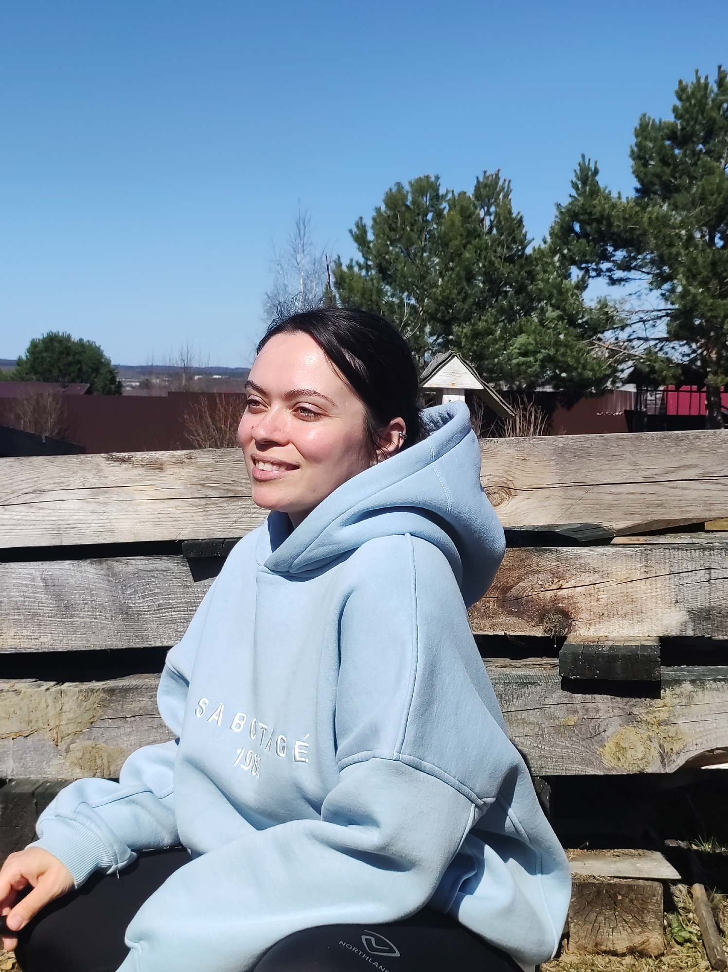 A young woman is sitting outdoors in Фоки, Russia, on a structure made of stacked wooden beams. She is wearing a light blue hoodie with the word "SABOTAGE" embroidered on the front, as well as black pants. The woman has dark hair pulled back and is smiling and looking to the side. In the background are trees and a fence, with a blue sky above. A birdhouse can be seen behind the woman. The setting appears to be rural or suburban.