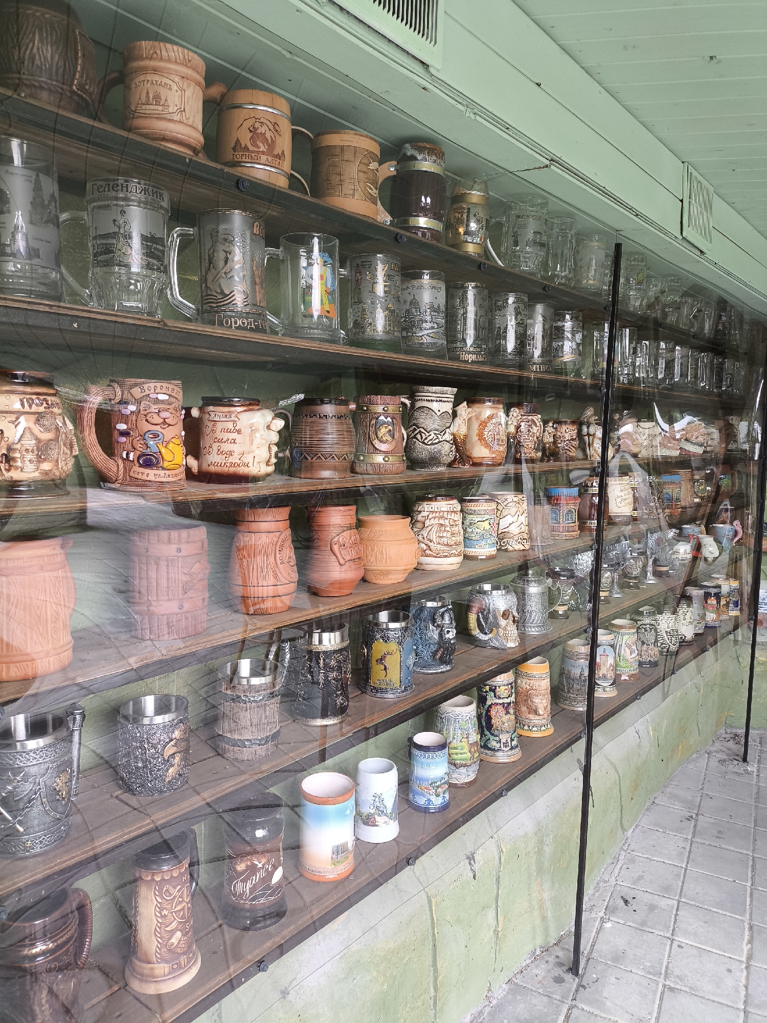 A display case filled with numerous beer mugs and other ceramic or glass drinking vessels is showcased in Borgustanskaya, Russia. The items are arranged on wooden shelves behind a clear protective covering, suggesting a shop or museum setting. The mugs vary in material, design, and size, with some featuring intricate carvings and painted scenes, while others are plain glass or metal. The backdrop of the display case is a light green wall, and the floor is paved with gray tiles. The lighting appears to be natural, filtering in from the front of the display.