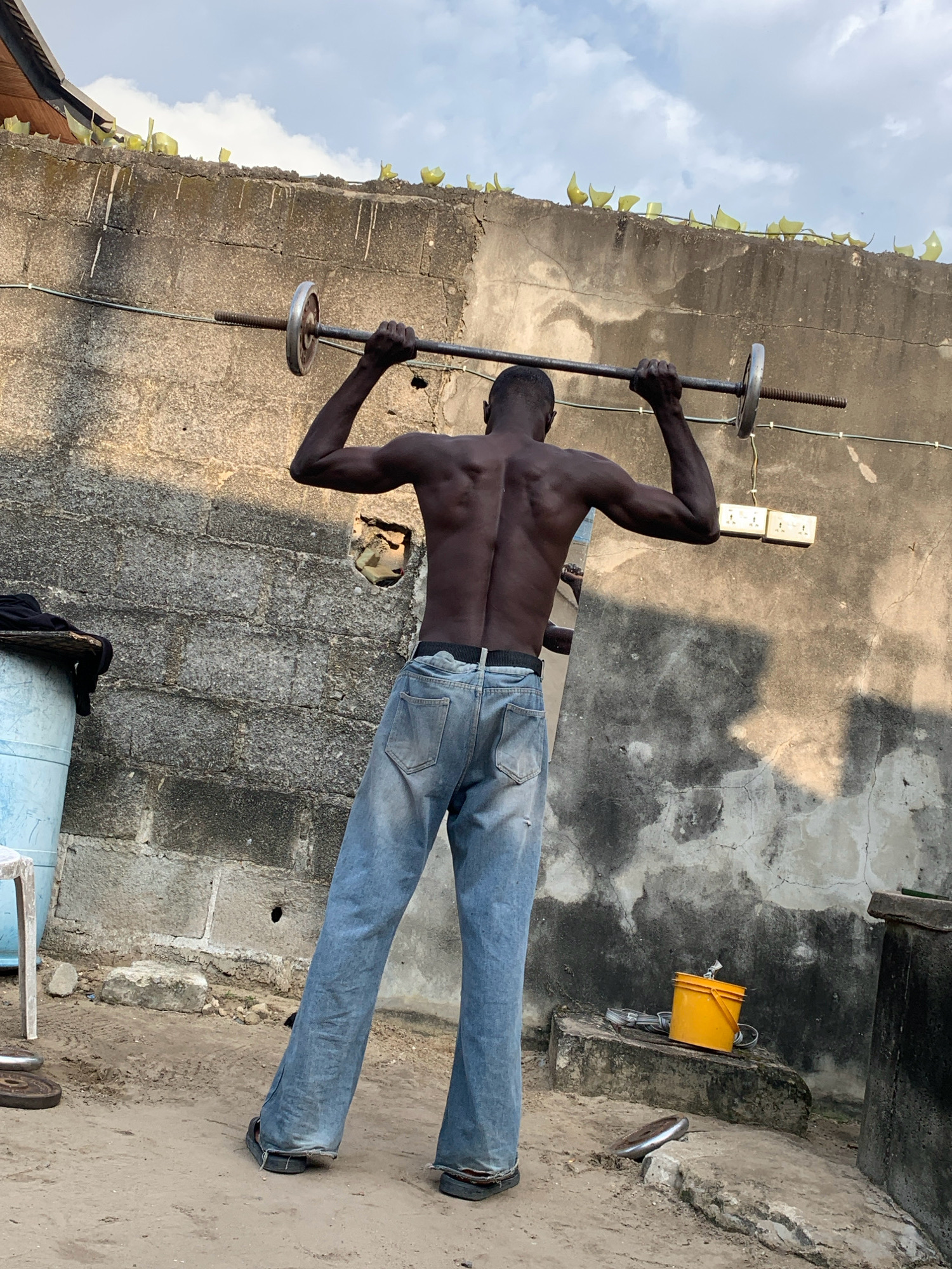 In Ado, Nigeria, a shirtless Black man, wearing light blue denim jeans and dark sandals, stands with his back to the camera, performing a barbell exercise. He is in an outdoor area with a rough concrete wall behind him, which has broken glass bottles embedded along its top edge. The wall has several visible electrical outlets. To the left of the man, a light blue plastic barrel is visible, with some black fabric draped over it. The ground is sandy and dirt. To the right, a bright yellow bucket and a concrete structure are present. The sky above is partly cloudy.