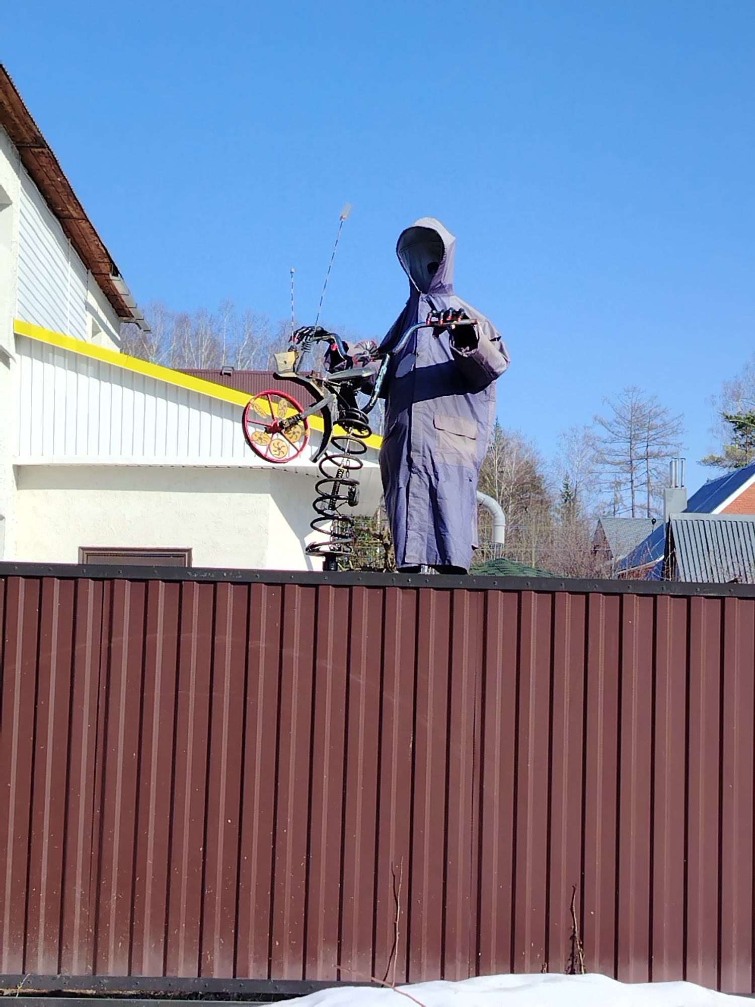 A humanoid figure draped in blue fabric stands atop a brown corrugated metal fence in Noviy, Russia. The figure wears a hood and holds what appears to be a motorcycle steering component in its hands. A sculpture of a whimsical vehicle, incorporating springs and a red and yellow wheel, is positioned to the figure's left. Behind the fence, there's a white building with a yellow trim. Bare trees and other residential structures are visible in the background, set against a clear blue sky. Snow is present at the base of the fence.