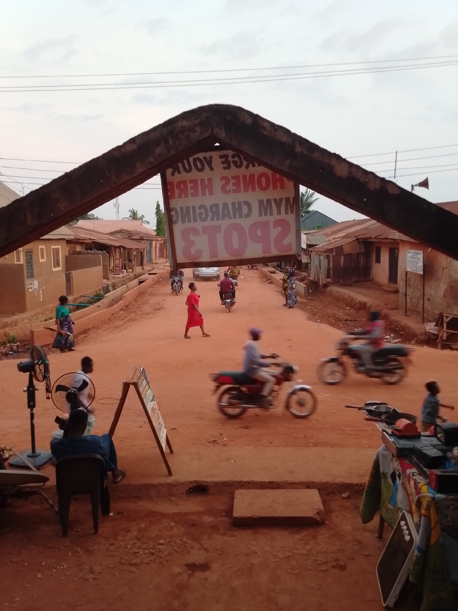 The image depicts a dusty street in Yelwa, Nigeria. Several people and motorcycles are moving along the road. A woman in a red dress is crossing the street. Buildings line both sides of the road. In the foreground, a person sits in a chair in front of two electric fans and a sandwich board. A table laden with items is also present. A sign with reversed text hangs over the street entrance: "SPOTS ARE HERE." "HOMES, PHONES, AND MYM CHARGING" are also listed.