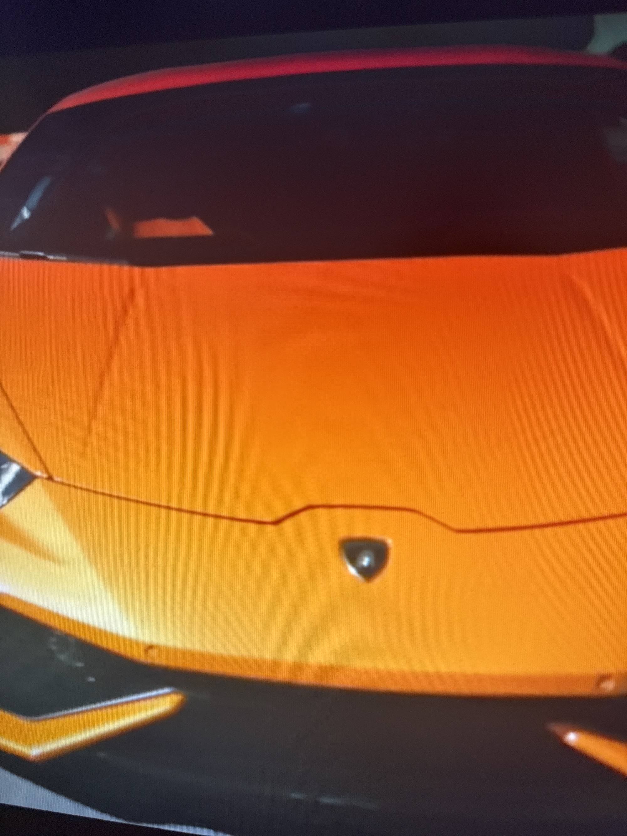 An orange Lamborghini sports car is parked in what appears to be an indoor parking garage, possibly in Netanya, Israel. The car's hood and headlights are visible. The lighting is dim, and the environment is dark, suggesting it is nighttime or an underground location. There are no people or other objects of interest visible in the frame.