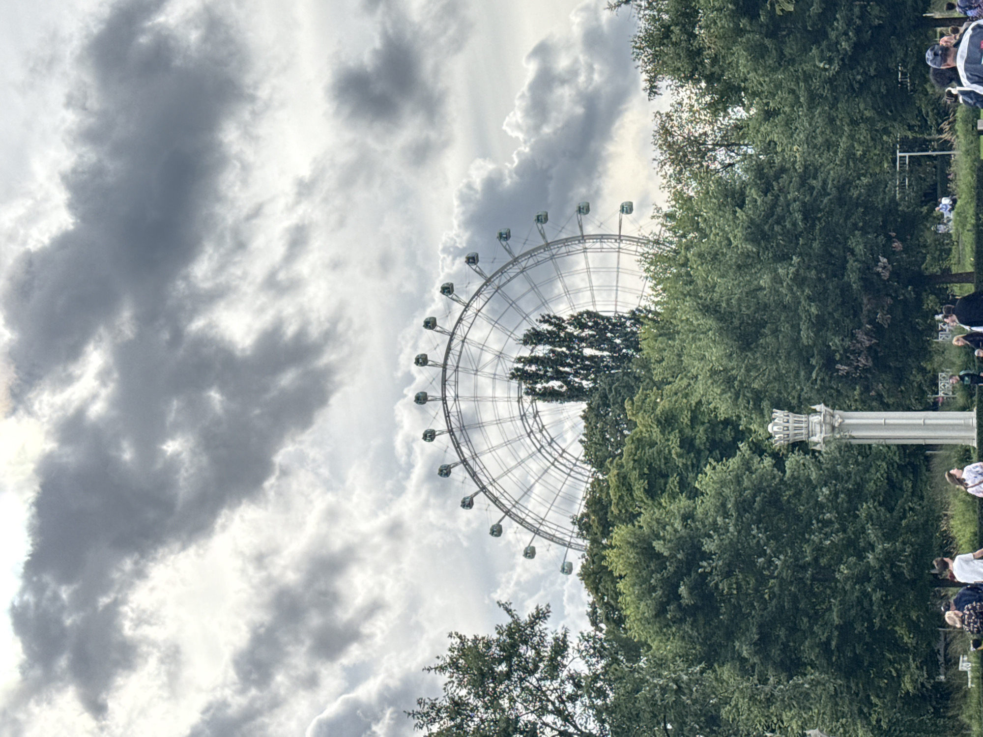 The image shows a Ferris wheel partially obscured by trees and dramatic clouds in Moscow, Russia. In the foreground, a white ornate pillar stands to the right of a group of people walking and standing. The Ferris wheel is a large, silver structure with numerous gondolas. The sky is filled with heavy, grey clouds, suggesting overcast weather. The trees in the middle ground are lush and green, forming a dense line. The overall scene depicts an outdoor recreational area with a mix of natural and man-made elements.