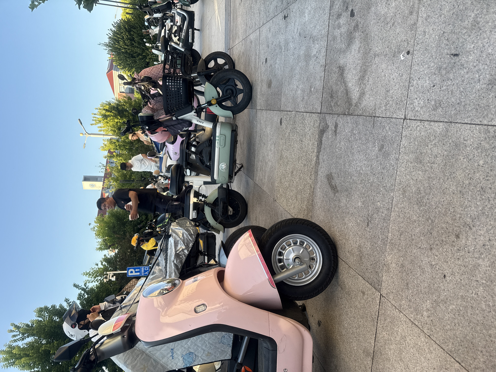 The image depicts a street scene in Qinhuangdao, China, with multiple electric scooters parked on a paved area. In the foreground, a light pink scooter is visible, with its wheel and part of its body clearly in frame. Behind it, several other scooters of various colors, including light green and white, are parked. A man in a black t-shirt is holding a cup and speaking to someone to his left, who is wearing a white helmet and black mask. Another person in a white t-shirt is standing near a light green scooter. Several trees and buildings are visible in the background, with a clear blue sky overhead. A blue and white parking sign is also present. The ground is made of light gray concrete tiles. The scene appears to be a public space, possibly a designated parking area for electric scooters.